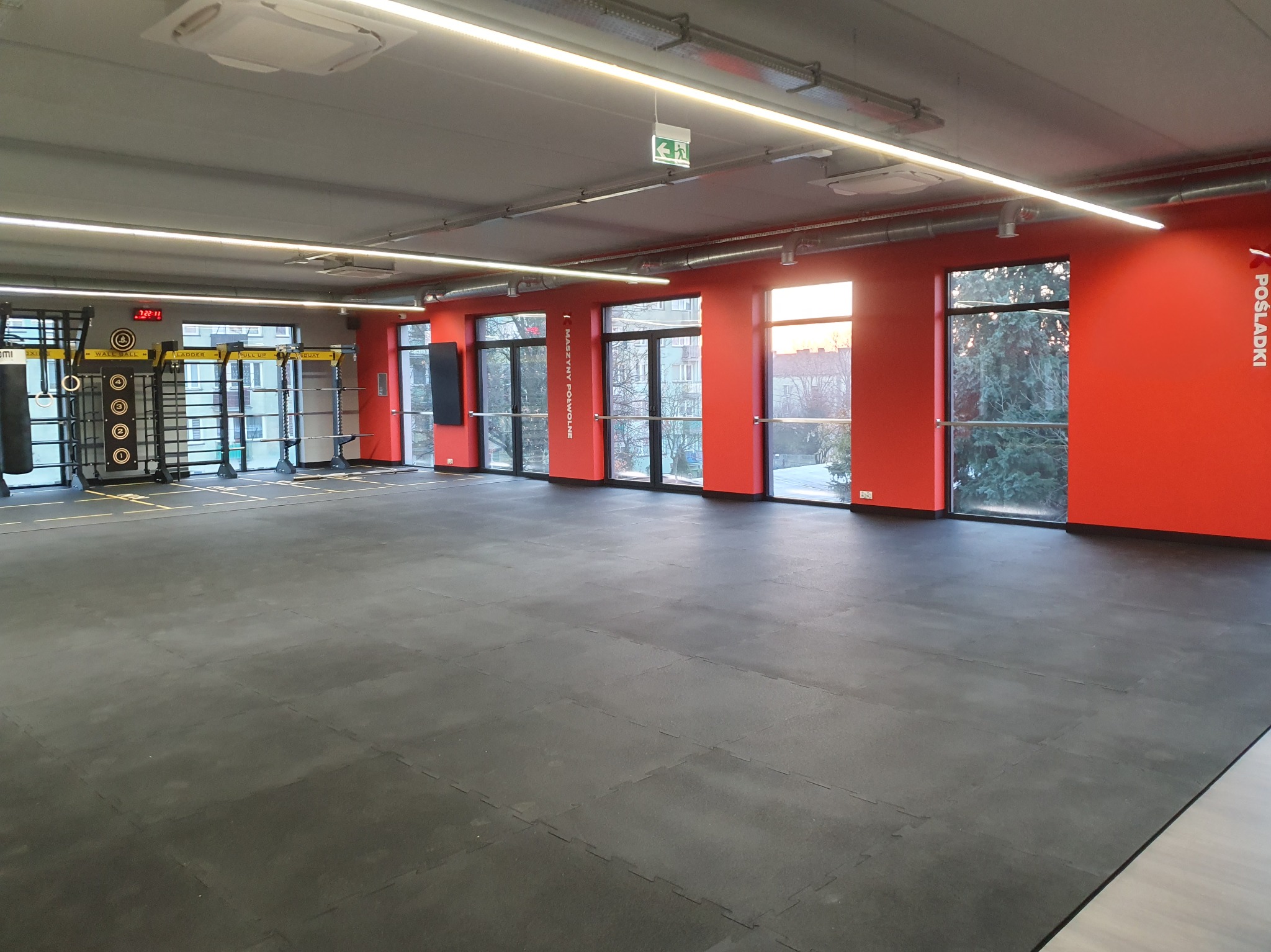 Nowoczesne wnętrze siłowni z czarną podłogą z gumowych puzzli, czerwonymi ścianami i dużymi oknami. Widoczne wyposażenie do ćwiczeń siłowych i bokserskich.