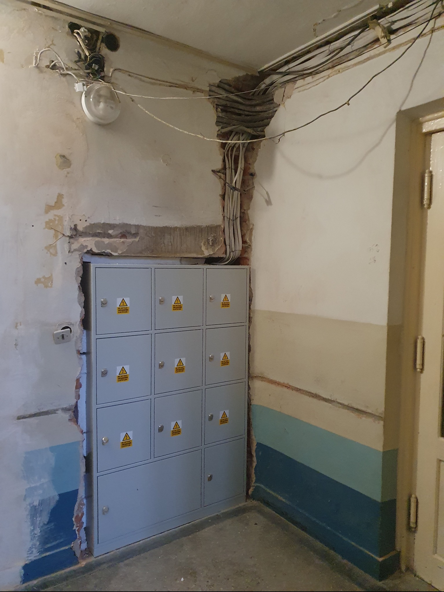Montaż szarej skrzynki elektrycznej z ostrzeżeniami w Łopusznie. Ściany w trakcie remontu, widoczne kable i instalacje elektryczne. Prace wykończeniowe w toku.