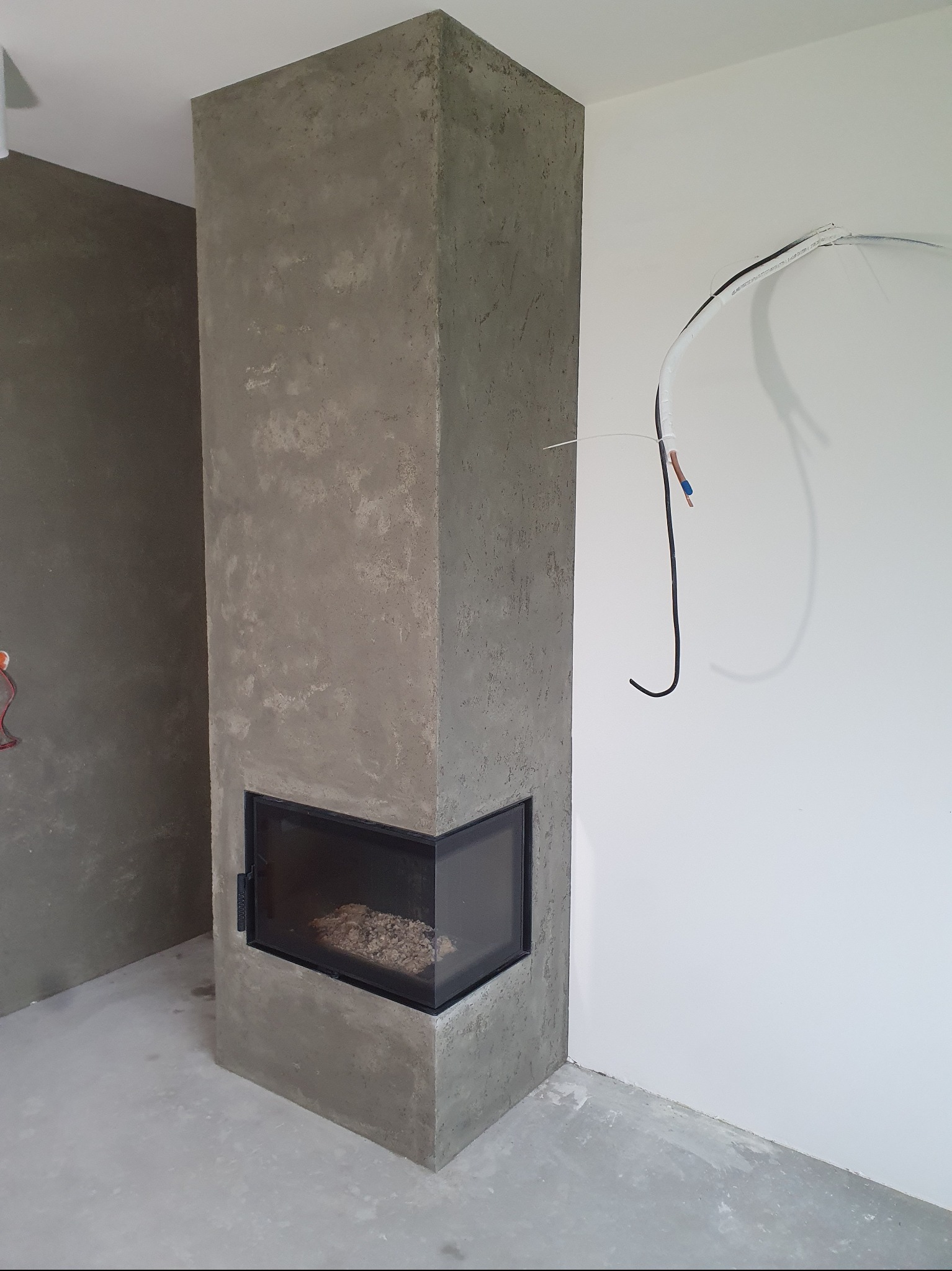 Wykończony kominek narożny z szarą, surową fakturą betonu, widoczny w nowym wnętrzu z instalacją elektryczną na ścianie.