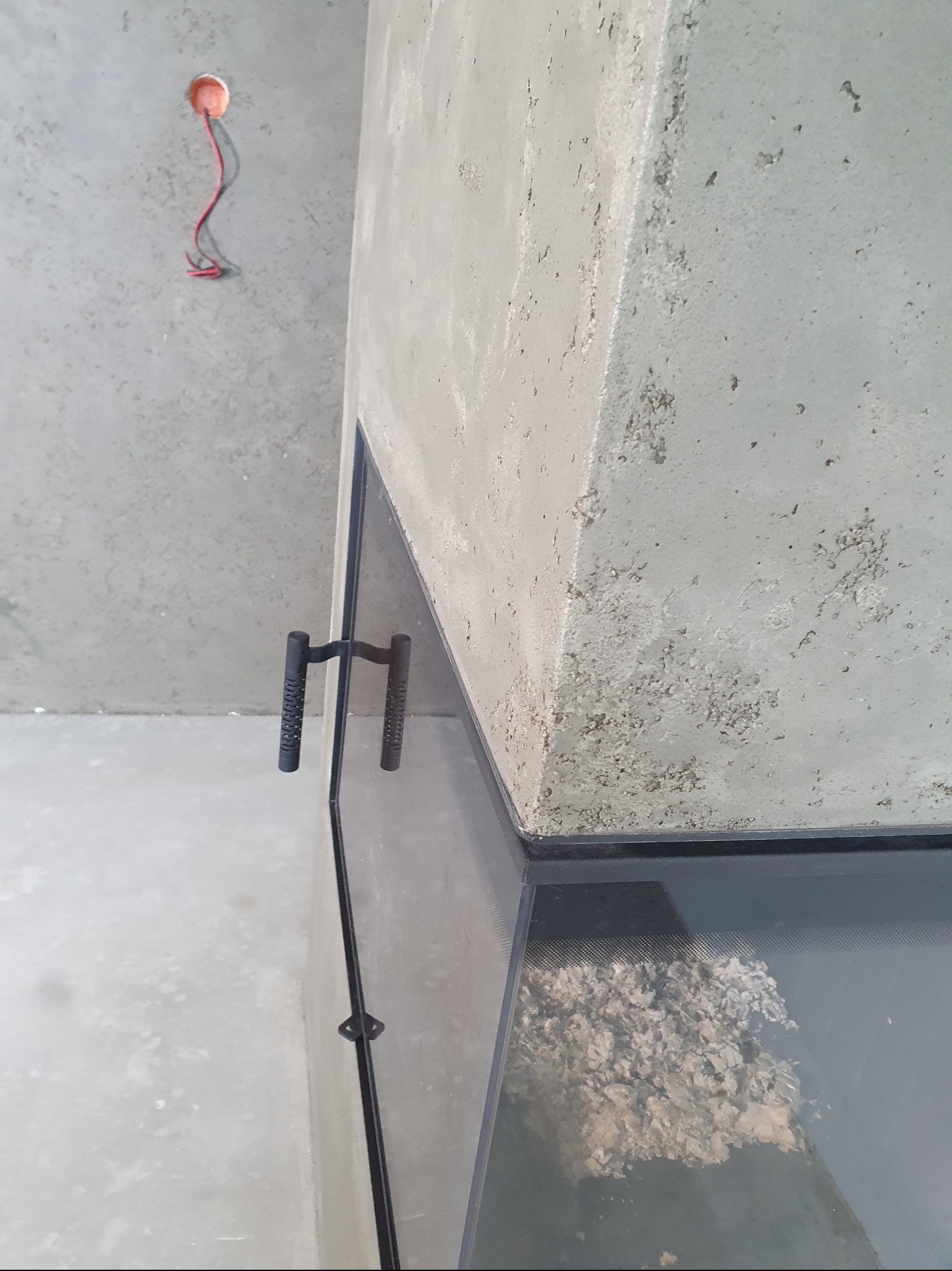 Surowy beton kominka z czarnymi uchwytami i szklaną szybą. W tle szara ściana z wystającymi kablami. Minimalistyczny design, surowe wykończenie.