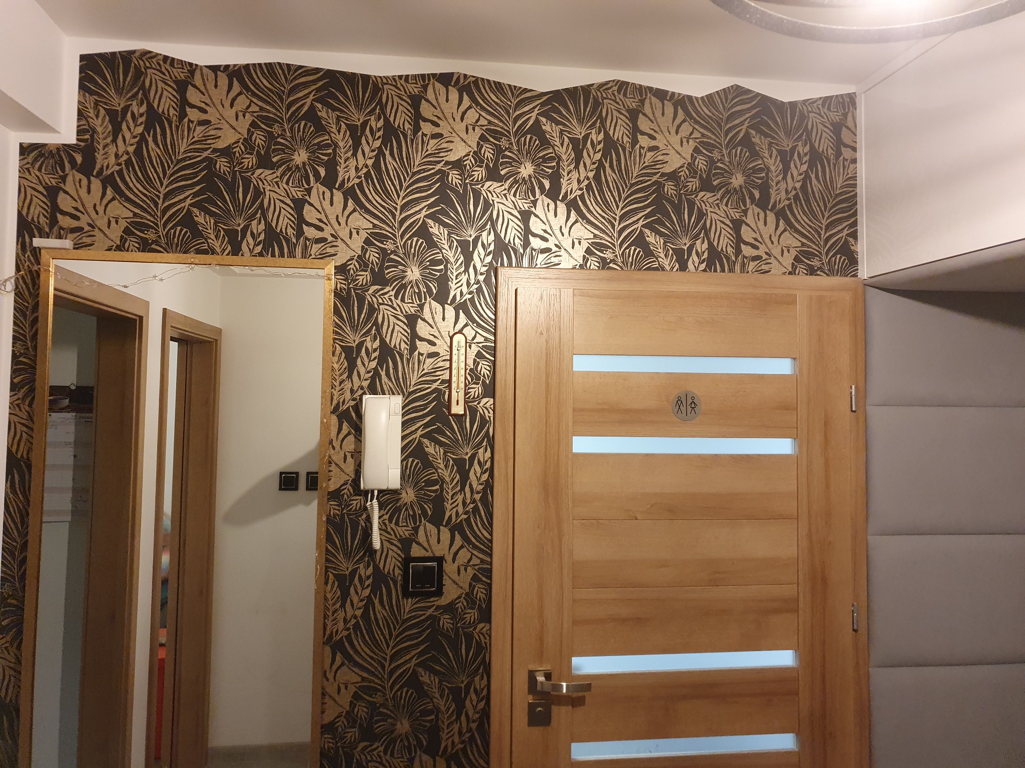 Wykończenie wnętrza: ściana z tapetą w liście, drewniane drzwi z przeszkleniami i dekoracyjna ściana obok, lustro w złotej ramie, domofon.