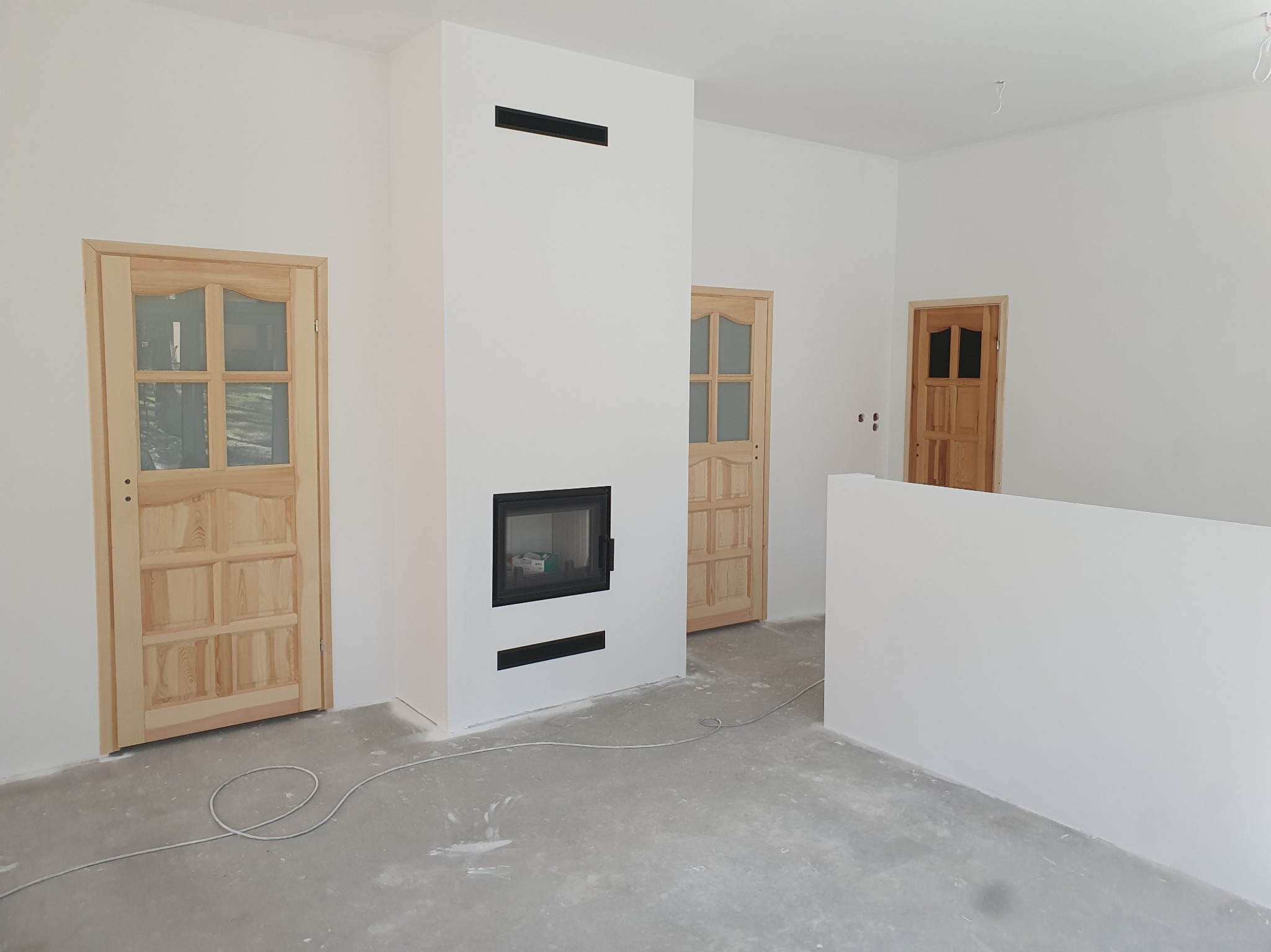 Jasne wnętrze z kominkiem obudowanym białą płytą, drewniane drzwi z przeszkleniami, surowa podłoga, świeżo pomalowane ściany. Minimalistyczny design.