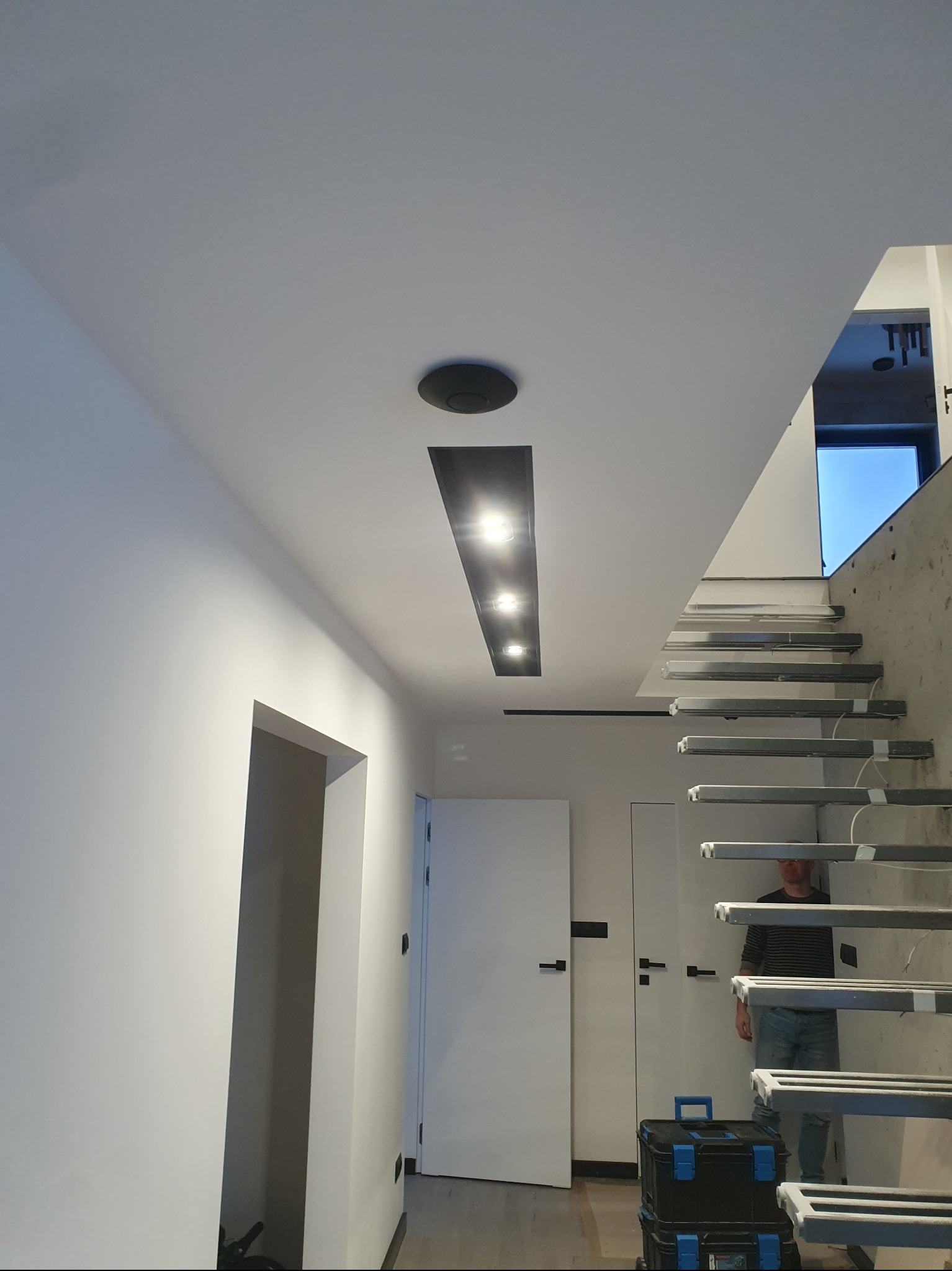 Nowoczesne wnętrze z białymi ścianami, oświetleniem sufitowym i schodami. Widoczny fragment osoby i skrzynki narzędziowe.