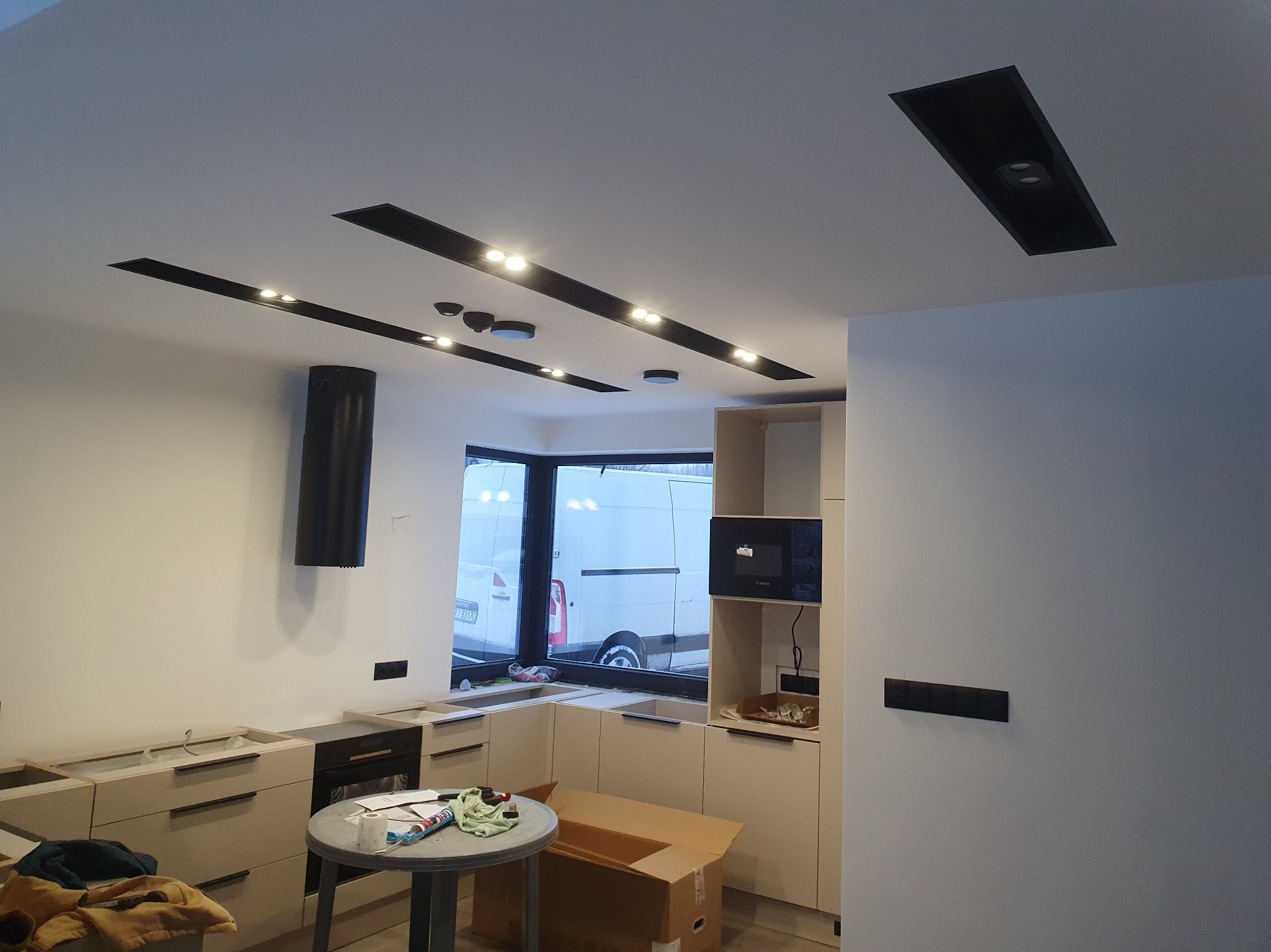 Nowoczesna kuchnia w trakcie montażu: jasne szafki, czarny okap i listwy oświetleniowe w suficie, duże okno z widokiem na zewnątrz. Widoczne elementy montażowe.