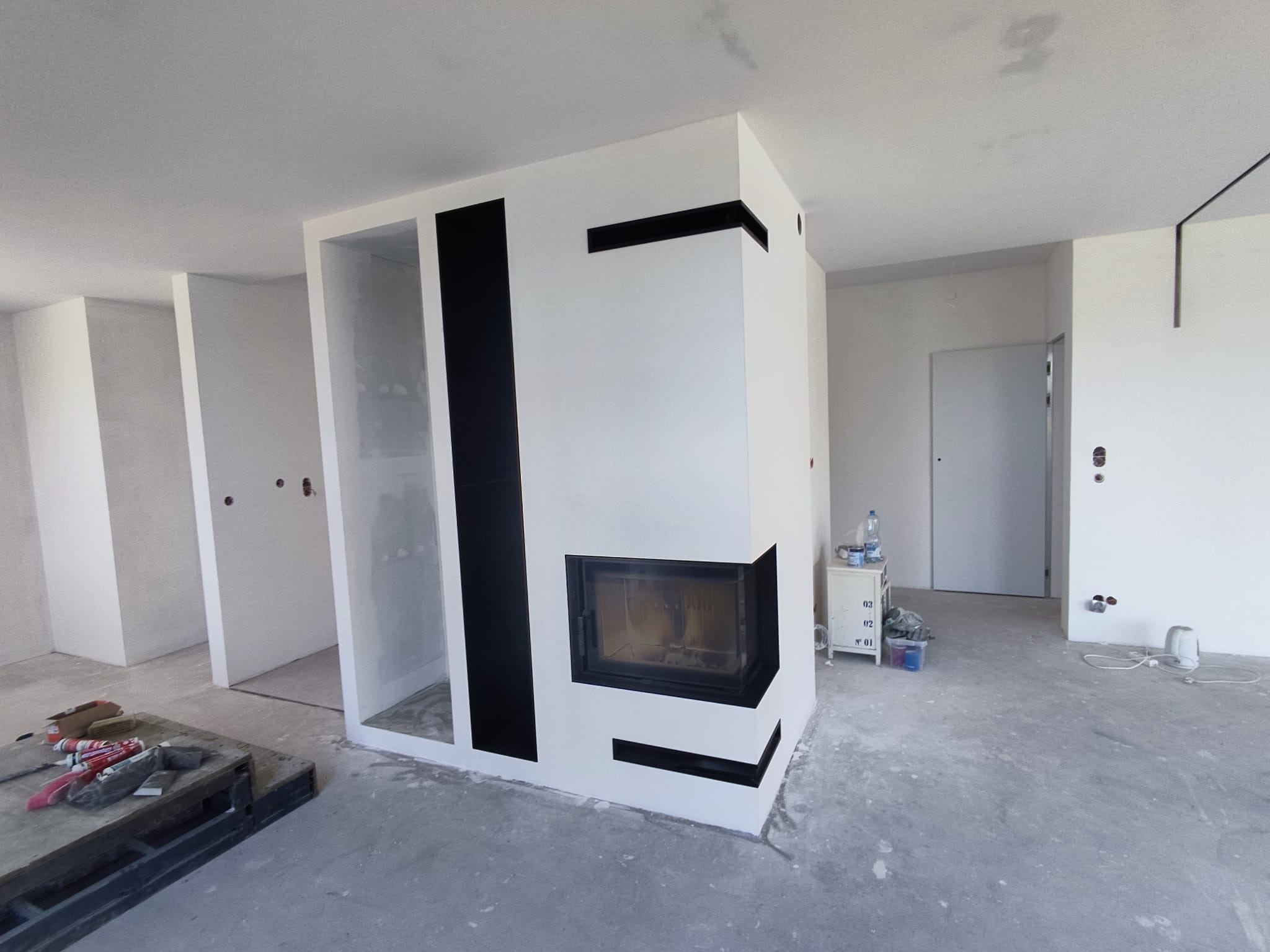 Nowoczesny kominek w zabudowie z czarnymi akcentami w surowym wnętrzu. Widoczne narzędzia malarskie sugerują etap wykończeniowy remontu.