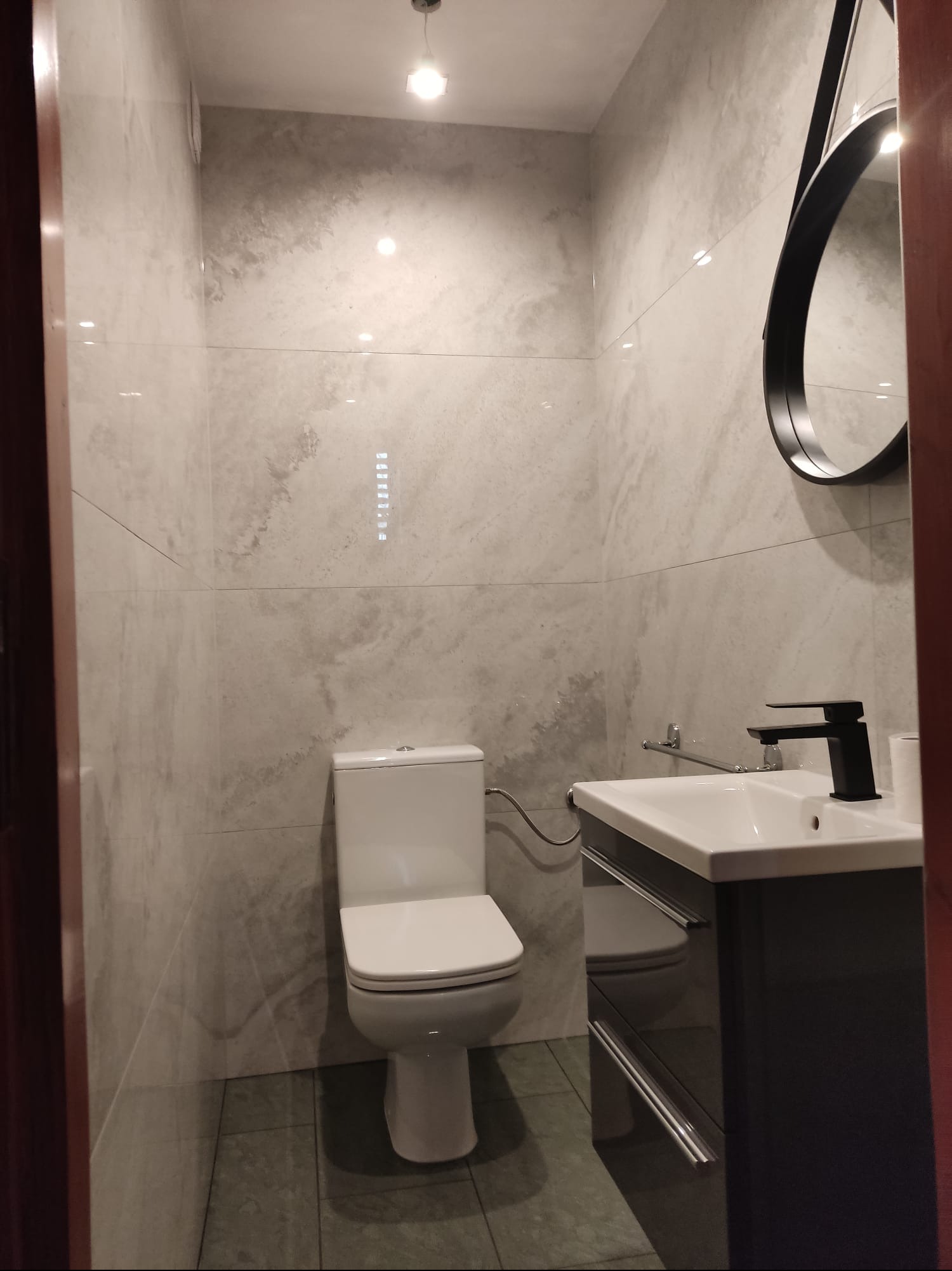 Wykończona łazienka z toaletą, umywalką z szafką i okrągłym lustrem. Ściany wyłożone jasnymi płytkami imitującymi kamień, szara podłoga. Nowoczesna armatura.