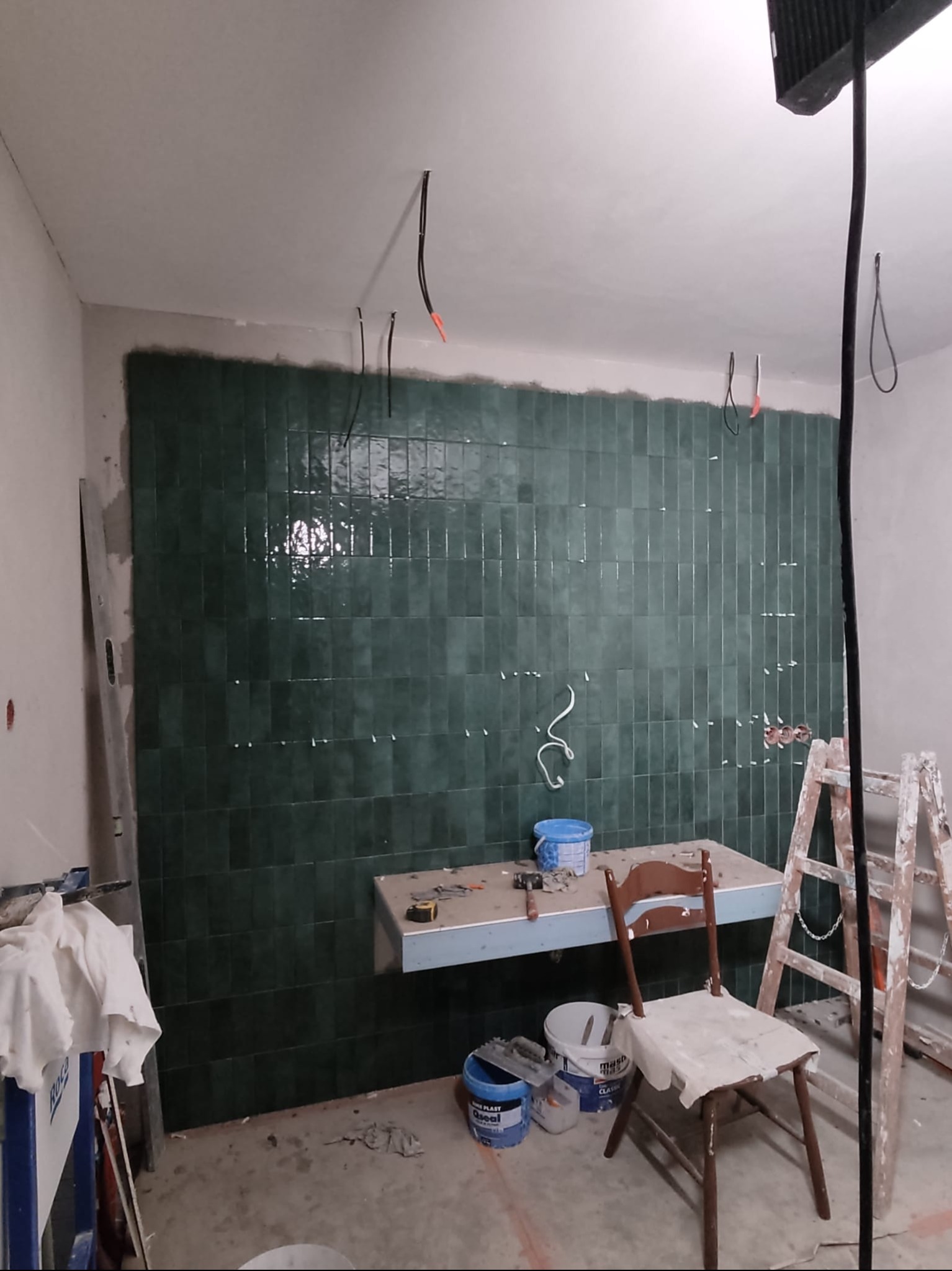 Ściana łazienki z połyskującymi, zielonymi płytkami ułożonymi w pionowe rzędy. Widoczne narzędzia, drabina i krzesło w trakcie prac wykończeniowych.