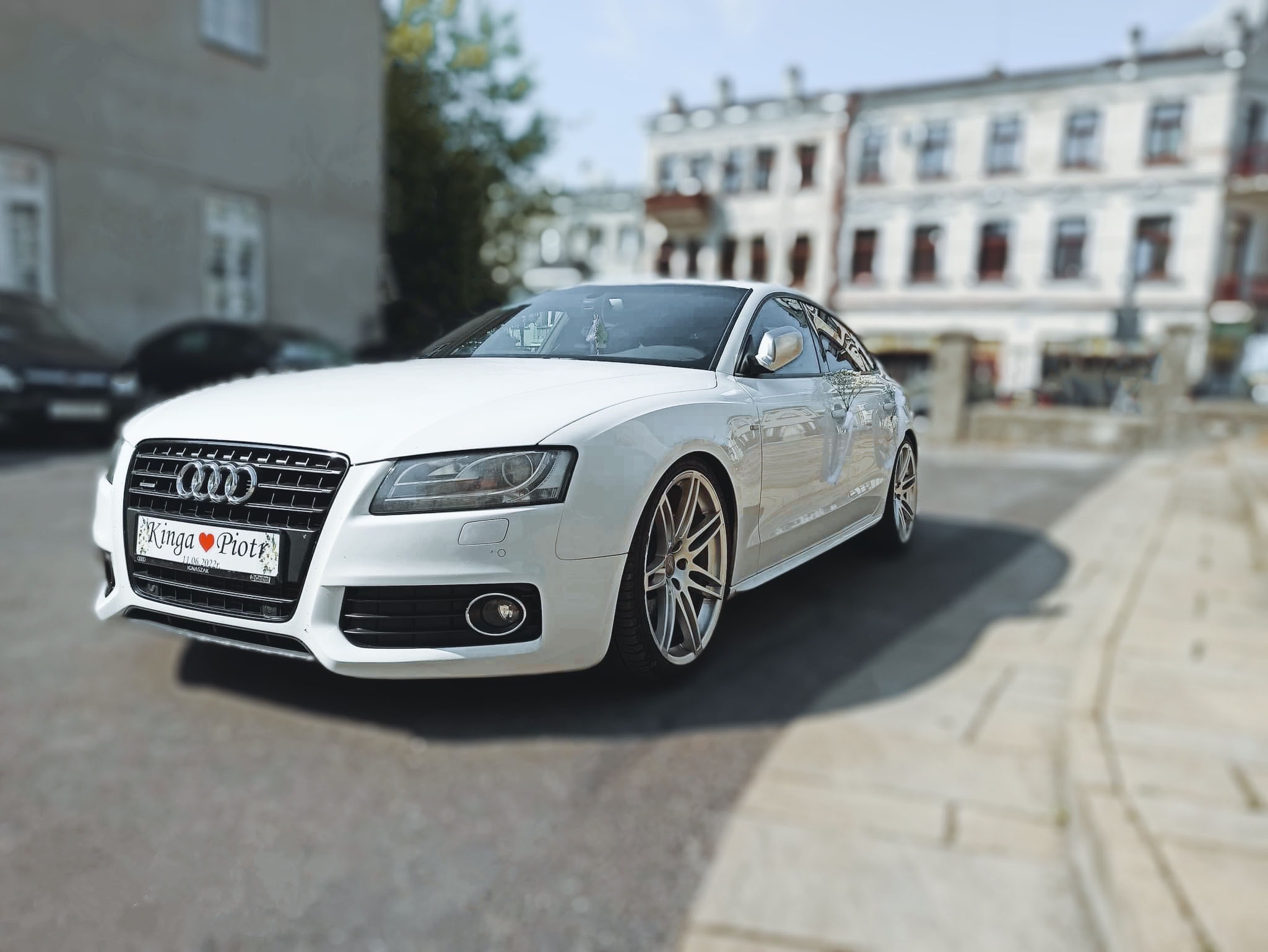 Białe Audi A5 z tablicą rejestracyjną 'Kinga Piotr' i datą ślubu, zaparkowane na tle miejskiej zabudowy.