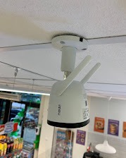 Biała kamera IP z dwiema antenami zamontowana na suficie w pomieszczeniu handlowym, widoczne półki z produktami w tle.