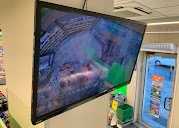 Ekran monitoringu CCTV w sklepie, widok z kamery na alejki z produktami, zamontowany na ścianie w rogu pomieszczenia, widoczny fragment sufitu i drzwi.