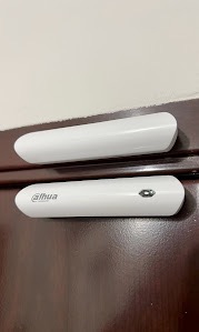 Dwa białe czujniki otwarcia drzwi/okien firmy Dahua zamontowane na ciemnobrązowych drzwiach, widoczny czujnik z logo i przyciskiem parowania.