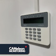 Klawiatura alarmowa z wyświetlaczem LCD marki Satel, zamontowana na białej ścianie, widoczny fragment logo firmy Cameleon Monitoring w lewym dolnym rogu.