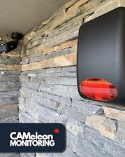 Zewnętrzny czujnik alarmowy z czerwoną diodą, zamontowany na ścianie z kamienia łupkowego, obok kamery monitoringu pod sufitem. Widoczne logo firmy CAMeleon MONITORING.