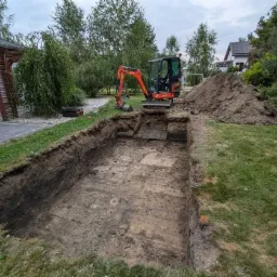 Wykonany wykop w ogrodzie, pomarańczowa mini koparka Kubota na skraju wykopu, pryzma ziemi obok, trawnik i zabudowa w tle.