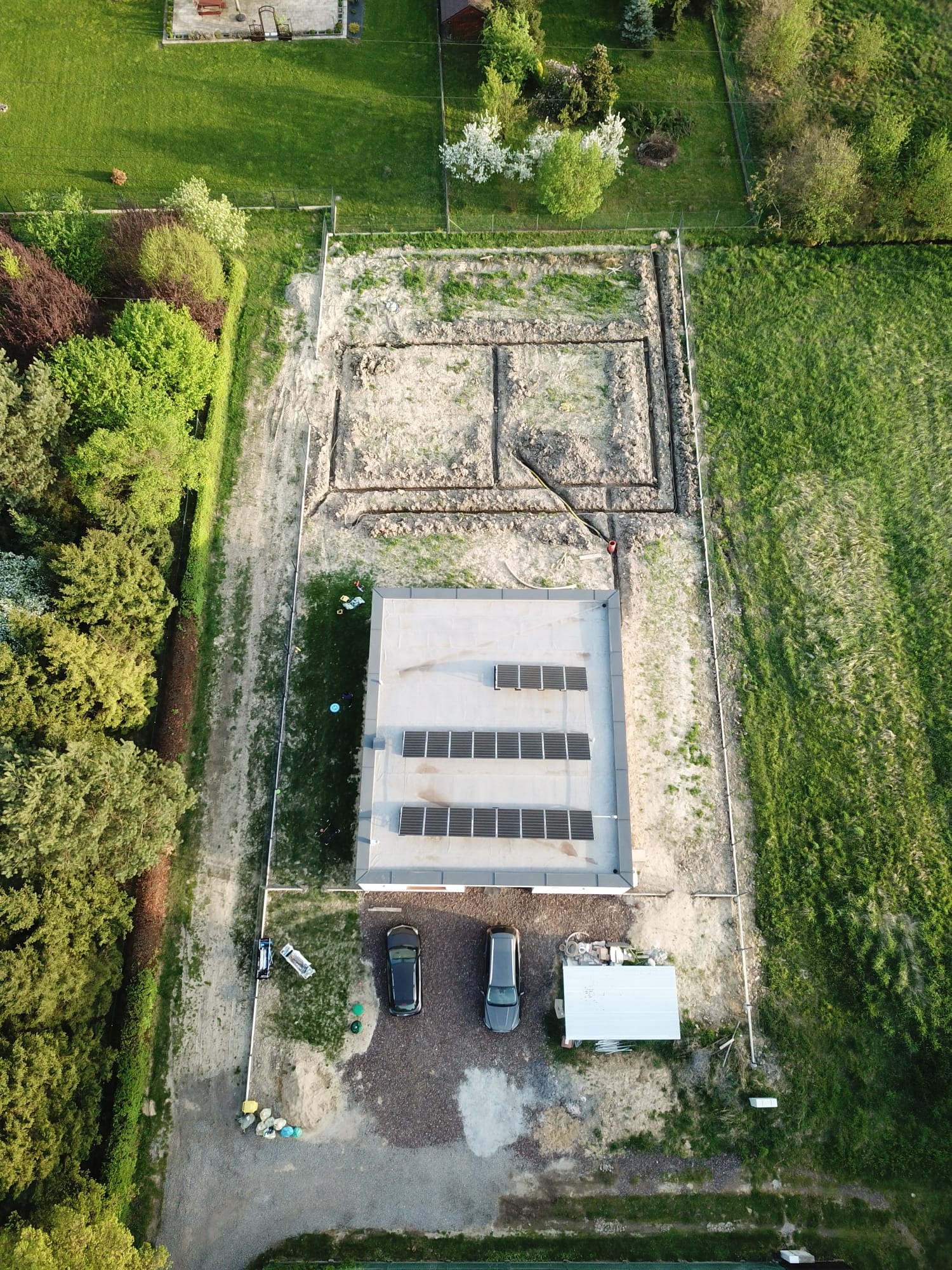 Widok z drona na plac budowy z wykopem pod fundamenty, obok budynek z panelami słonecznymi, dwa samochody osobowe i teren częściowo przygotowany pod trawnik.