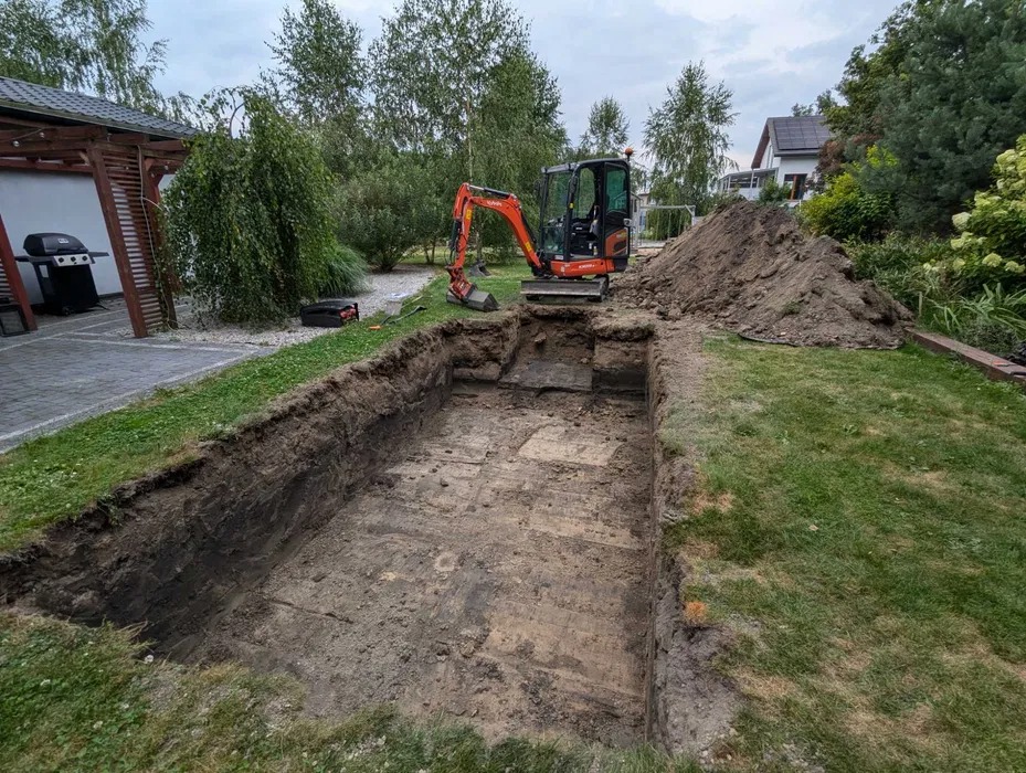 Wykonany wykop w ogrodzie, pomarańczowa mini koparka Kubota na skraju wykopu, pryzma ziemi obok, trawnik i zabudowa w tle.