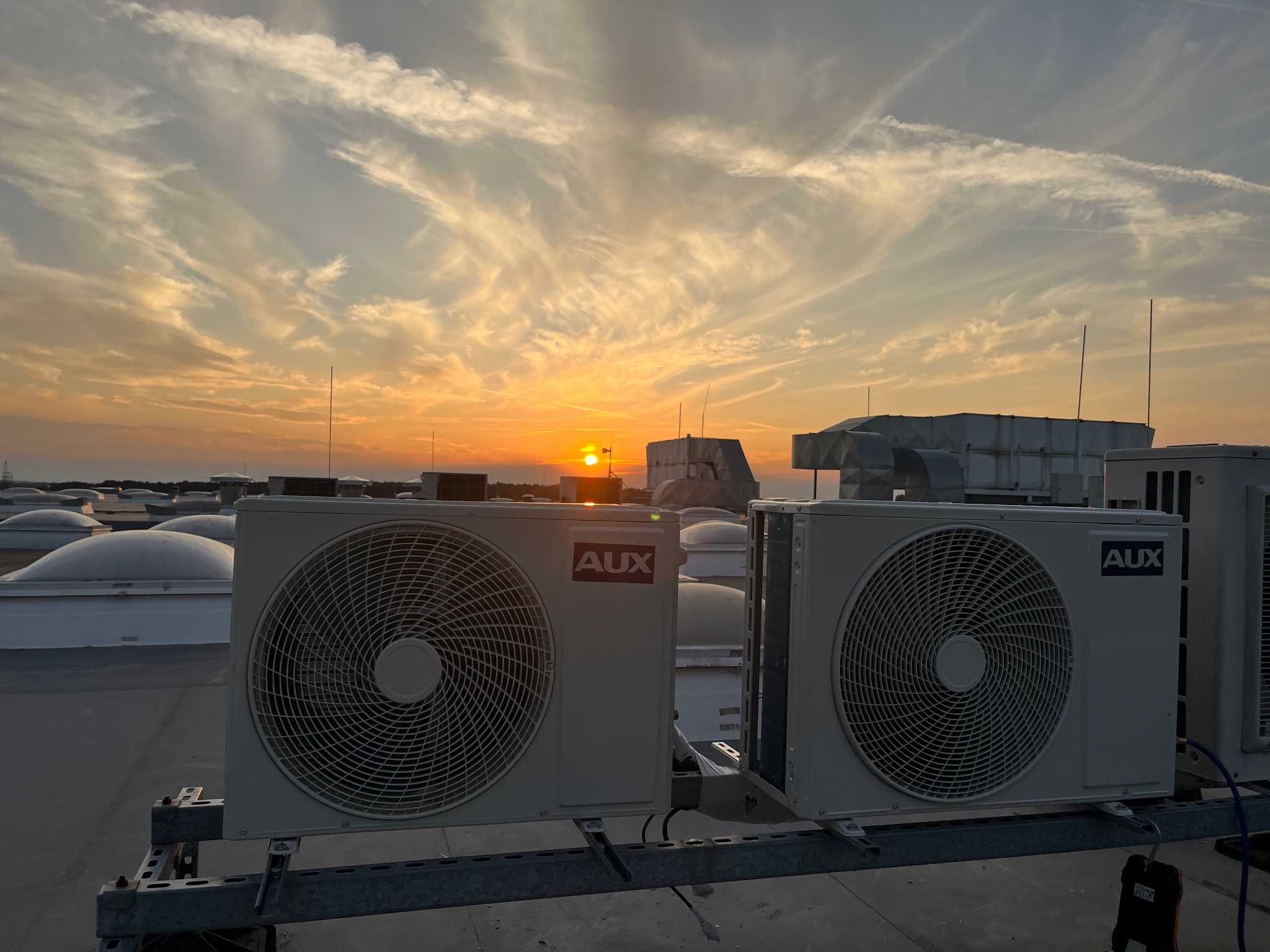 Zewnętrzne jednostki klimatyzacji AUX na dachu budynku o zmierzchu, z widocznym zachodzącym słońcem i obłokami na niebie.