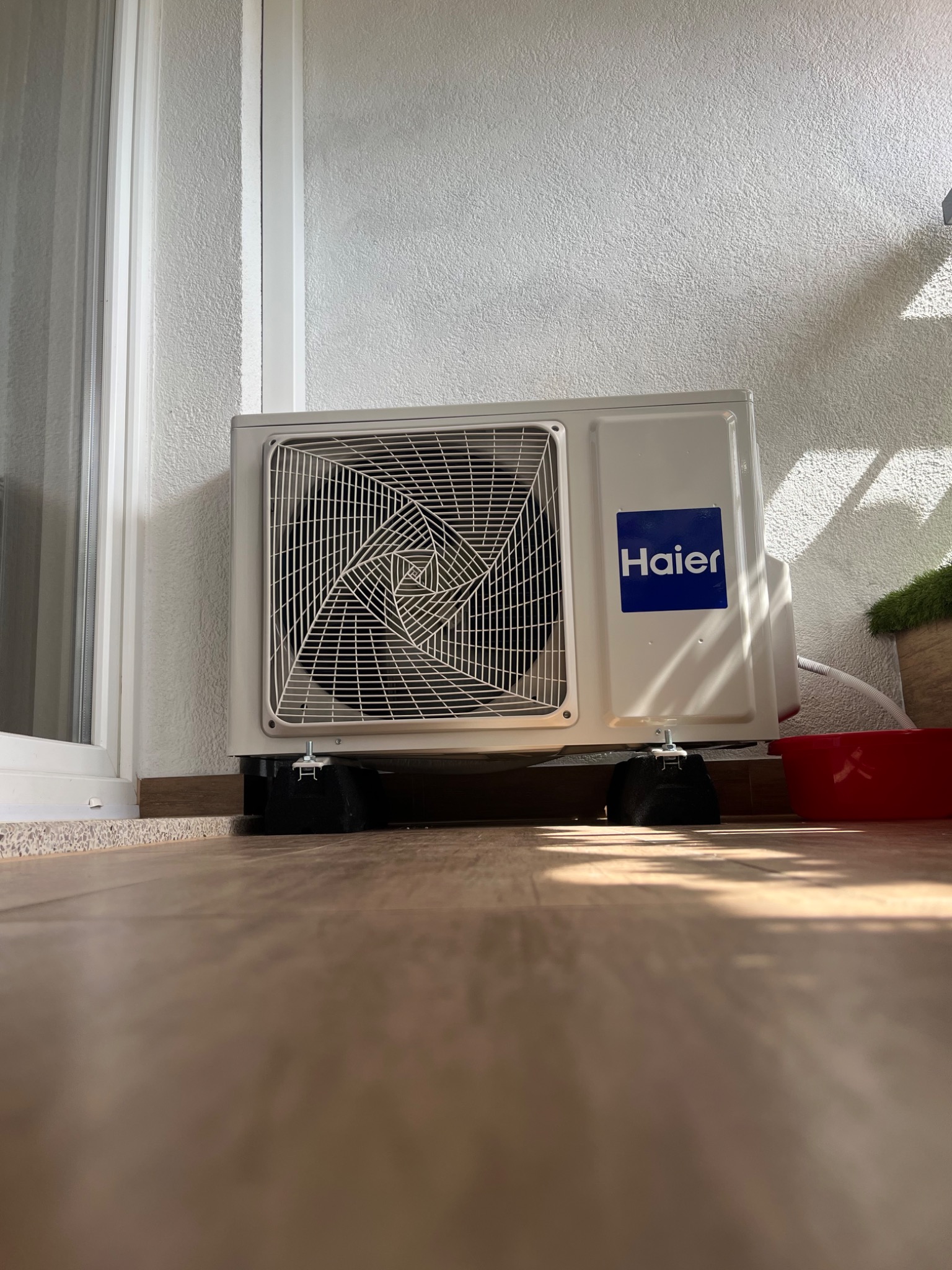 Biała jednostka zewnętrzna klimatyzacji Haier umieszczona na czarnych podstawkach antywibracyjnych na balkonie, widoczna czerwona miska i doniczka z zieloną rośliną.