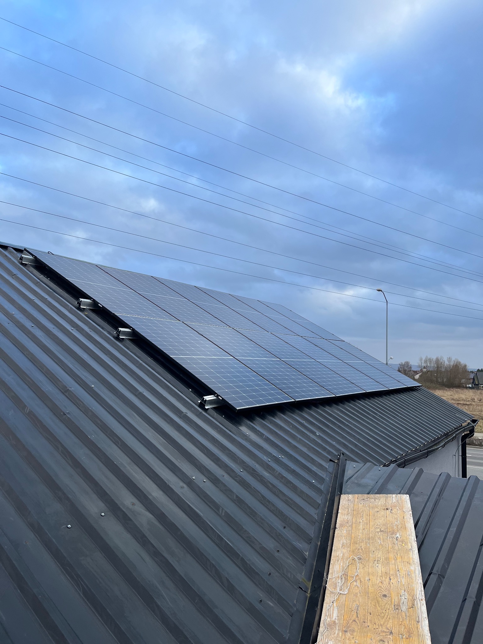 Panel słoneczny zamontowany na dachu z blachy trapezowej, widok z dołu na pochmurne niebo i linie energetyczne.