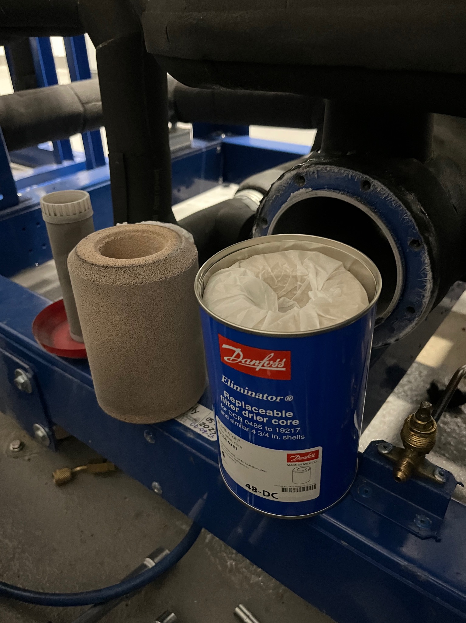 Filtr odwadniający Danfoss Eliminator w niebieskiej puszce obok szarego cylindrycznego wkładu na tle instalacji klimatyzacyjnej z rurami.