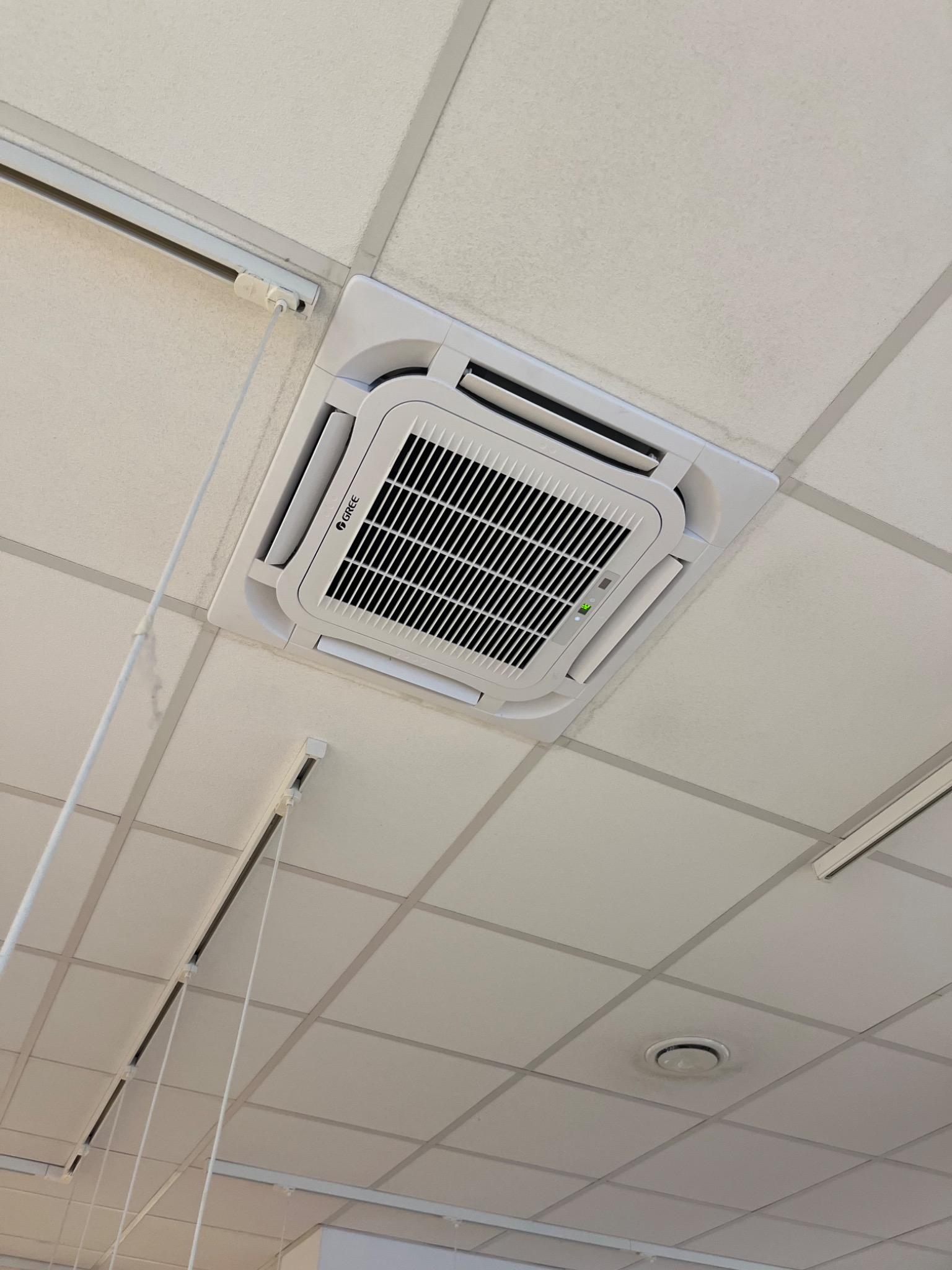 Kasetonowa jednostka klimatyzacji marki Gree zamontowana w suficie podwieszanym, widoczna zielona dioda sygnalizacyjna pracy urządzenia.