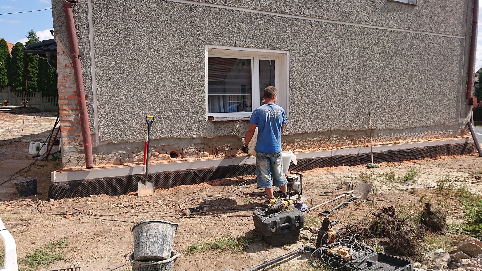 Prace przygotowawcze do izolacji fundamentów domu jednorodzinnego: widoczne odkopane fundamenty, zabezpieczone folią kubełkową, z pracownikiem w trakcie wykonywania prac, narzędzia budowlane...