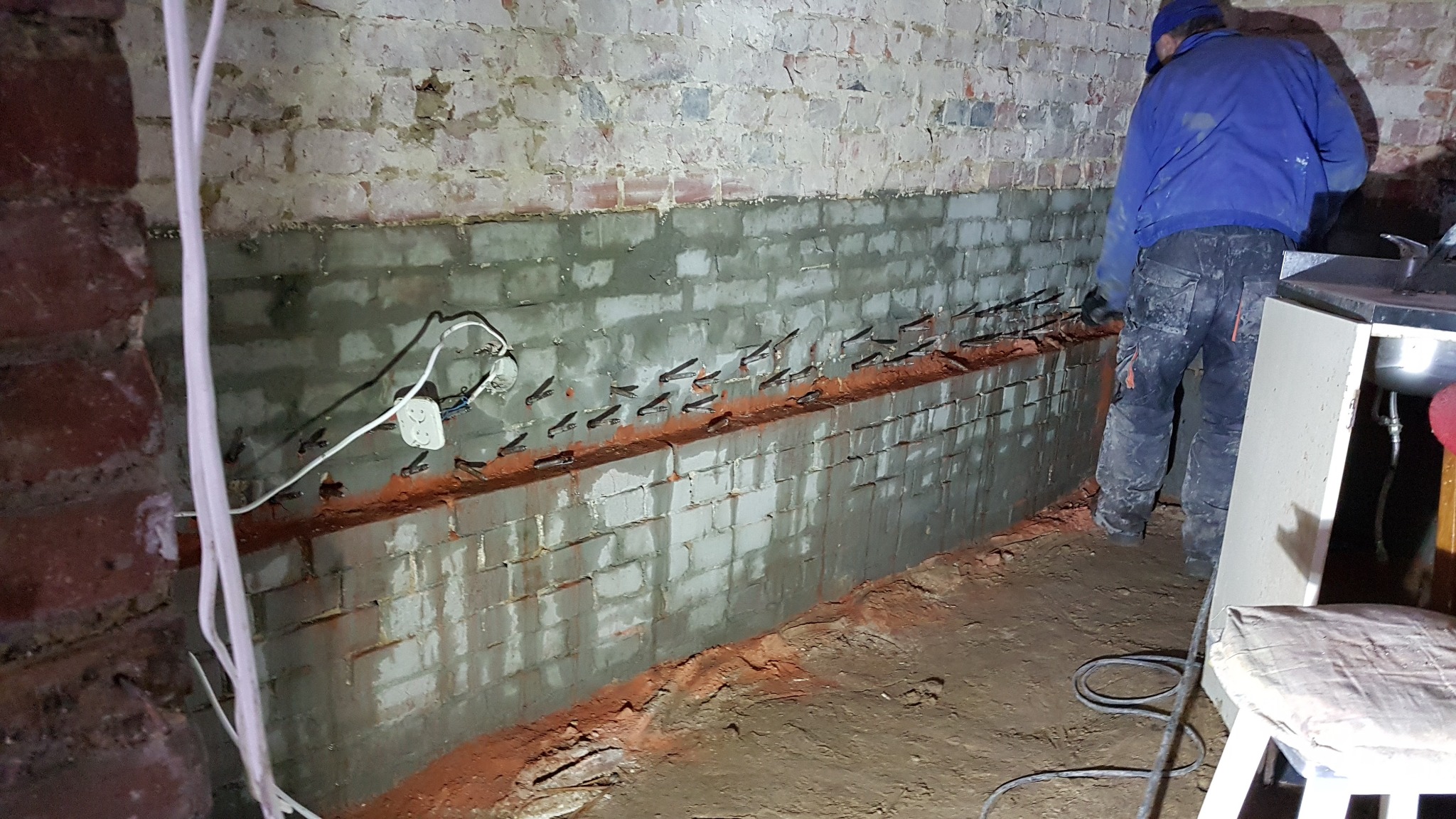 Prace przygotowawcze do izolacji pionowej fundamentu z widocznymi kotwami mocującymi, śladami wilgoci na cegłach i pracownikiem w roboczym ubraniu.