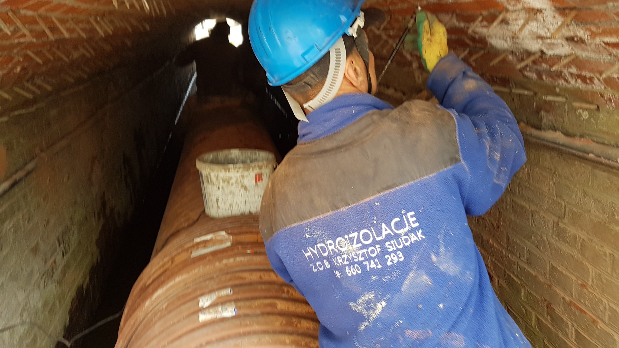 Pracownik w niebieskim kasku i bluzie z napisem 'HYDROIZOLACJE' aplikuje preparat na ceglany mur wewnątrz tunelu, obok stoi wiadro z resztkami materiału.