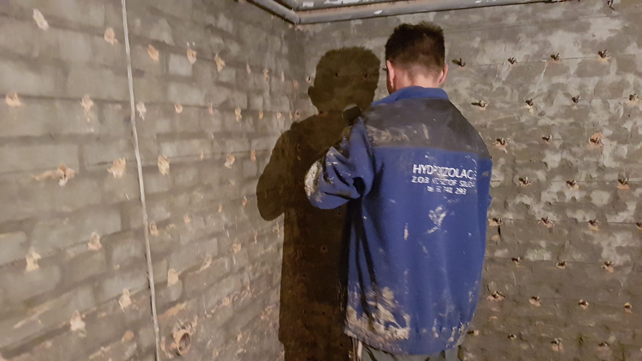 Pracownik w niebieskiej bluzie z napisem 'HYDROIZOLACJE' stoi tyłem do kamery na tle ściany fundamentowej po iniekcji ciśnieniowej, widoczne liczne otwory wypełnione szarym materiałem.