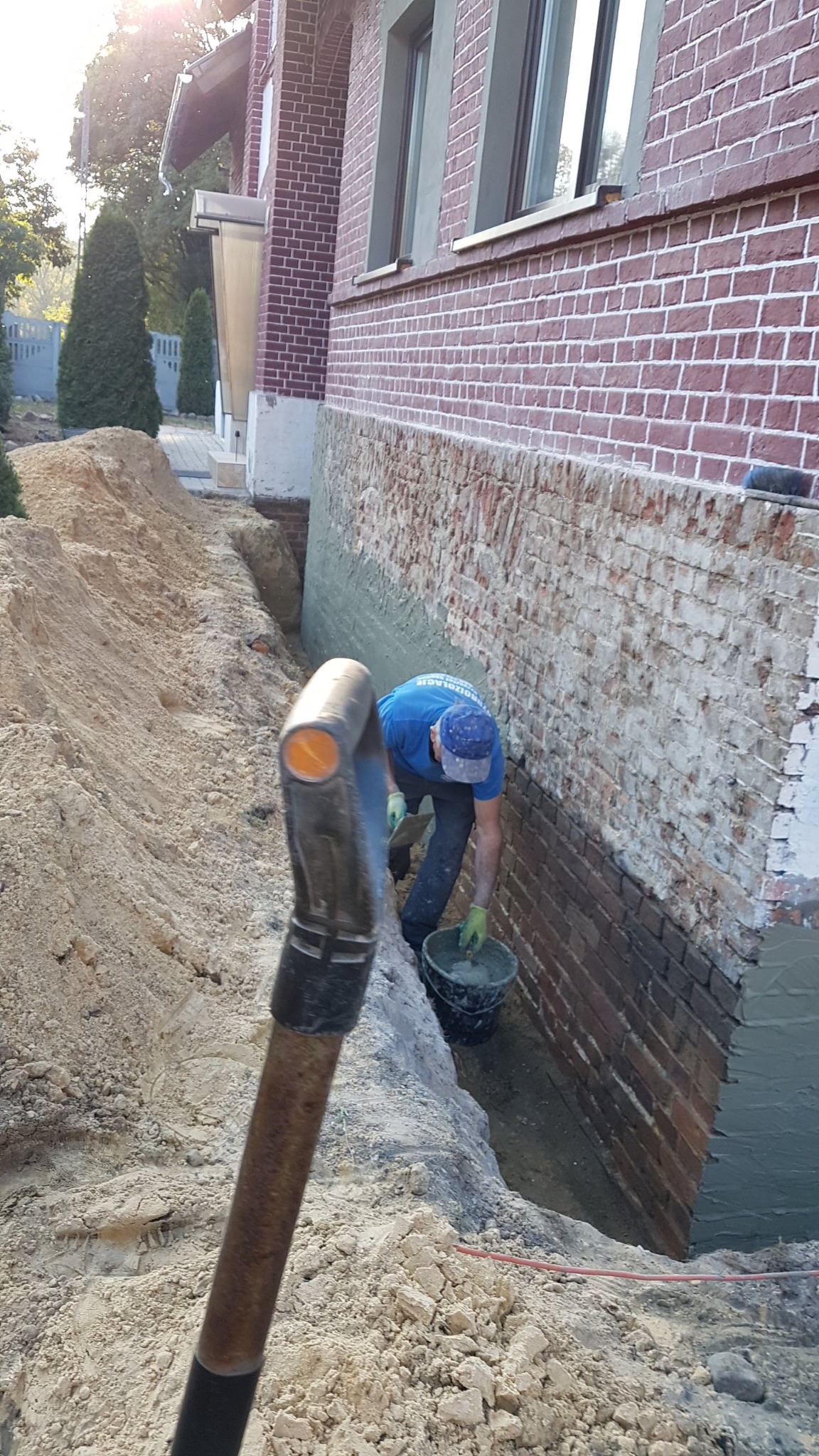 Pracownik w niebieskiej koszulce i czapce nanosi szarą zaprawę na ceglany fundament budynku, widoczne wykopane obok fundamentu podłoże i fragment narzędzia w pierwszym planie.