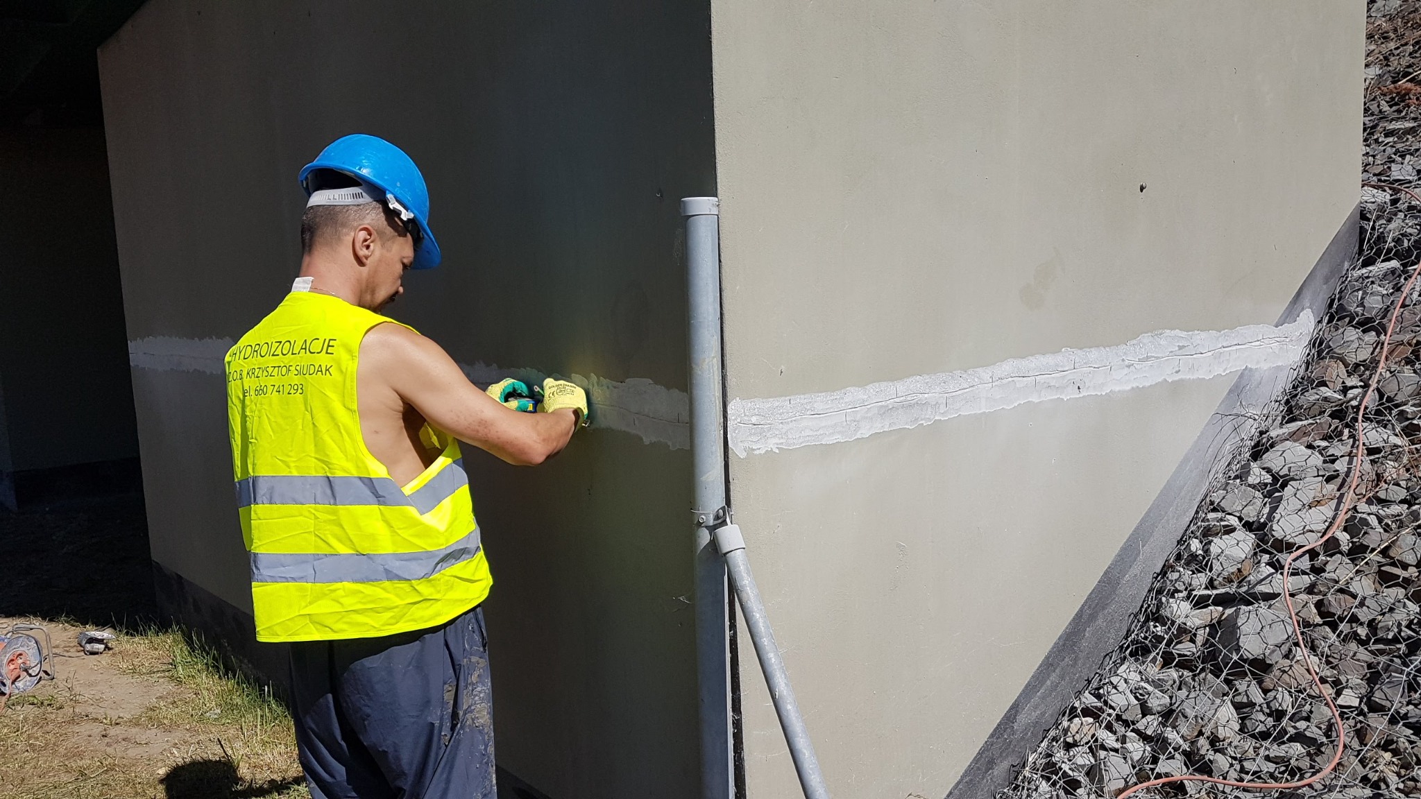 Pracownik w kamizelce odblaskowej i niebieskim kasku uszczelnia pęknięcie na fundamencie budynku, widoczne zabezpieczenie z kamieni w siatce.
