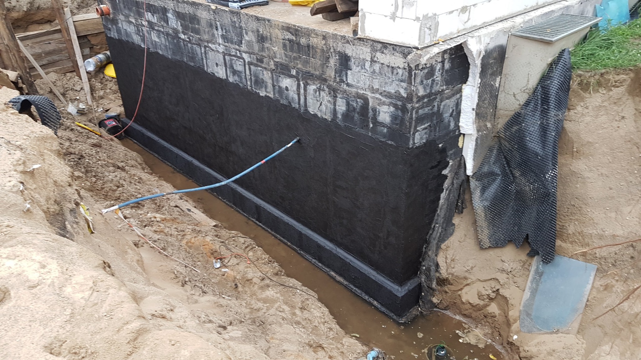Hydroizolacja pionowa fundamentu z użyciem czarnej masy bitumicznej, widoczna folia kubełkowa i drenaż w wykopie.