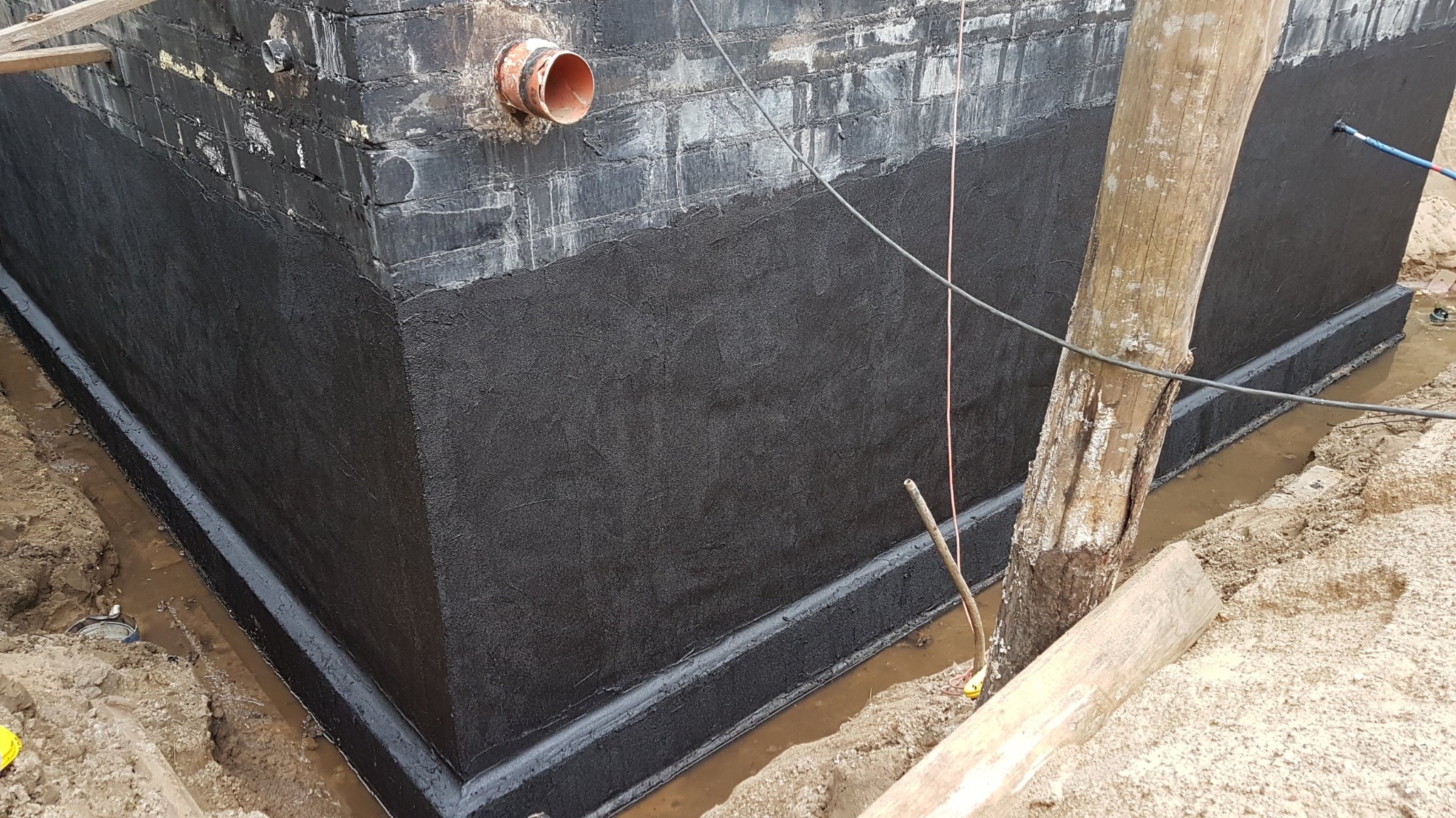 Ceglany fundament budynku z nałożoną czarną hydroizolacją bitumiczną, widoczna rura odpływowa i drewniany stempel podtrzymujący.