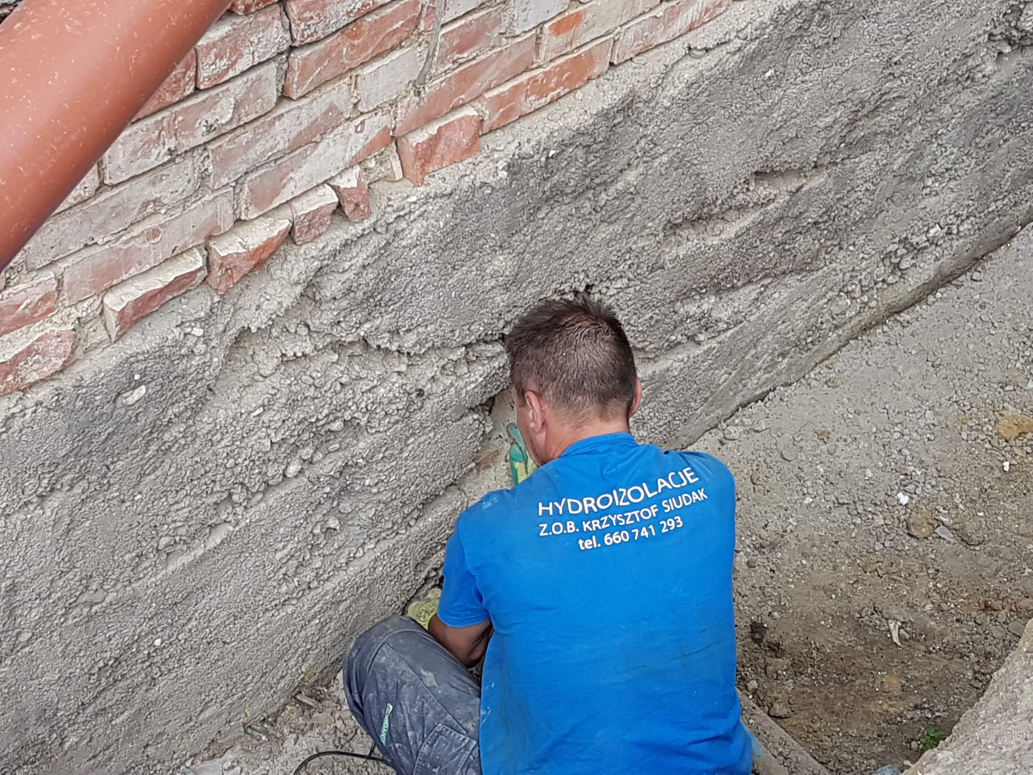 Pracownik w niebieskiej koszulce z napisem HYDROIZOLACJE Z.O.B. KRZYSZTOF SIUDAK, wykonuje uszczelnianie fundamentu przy użyciu narzędzia, widoczne cegły i świeży beton.
