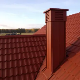 Czerwony dach z blachodachówki z kominem wentylacyjnym w tym samym kolorze, na tle zieleni drzew i błękitnego nieba.
