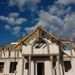 Surowy stan domu jednorodzinnego z widoczną konstrukcją drewnianą dachu na tle błękitnego nieba z chmurami.