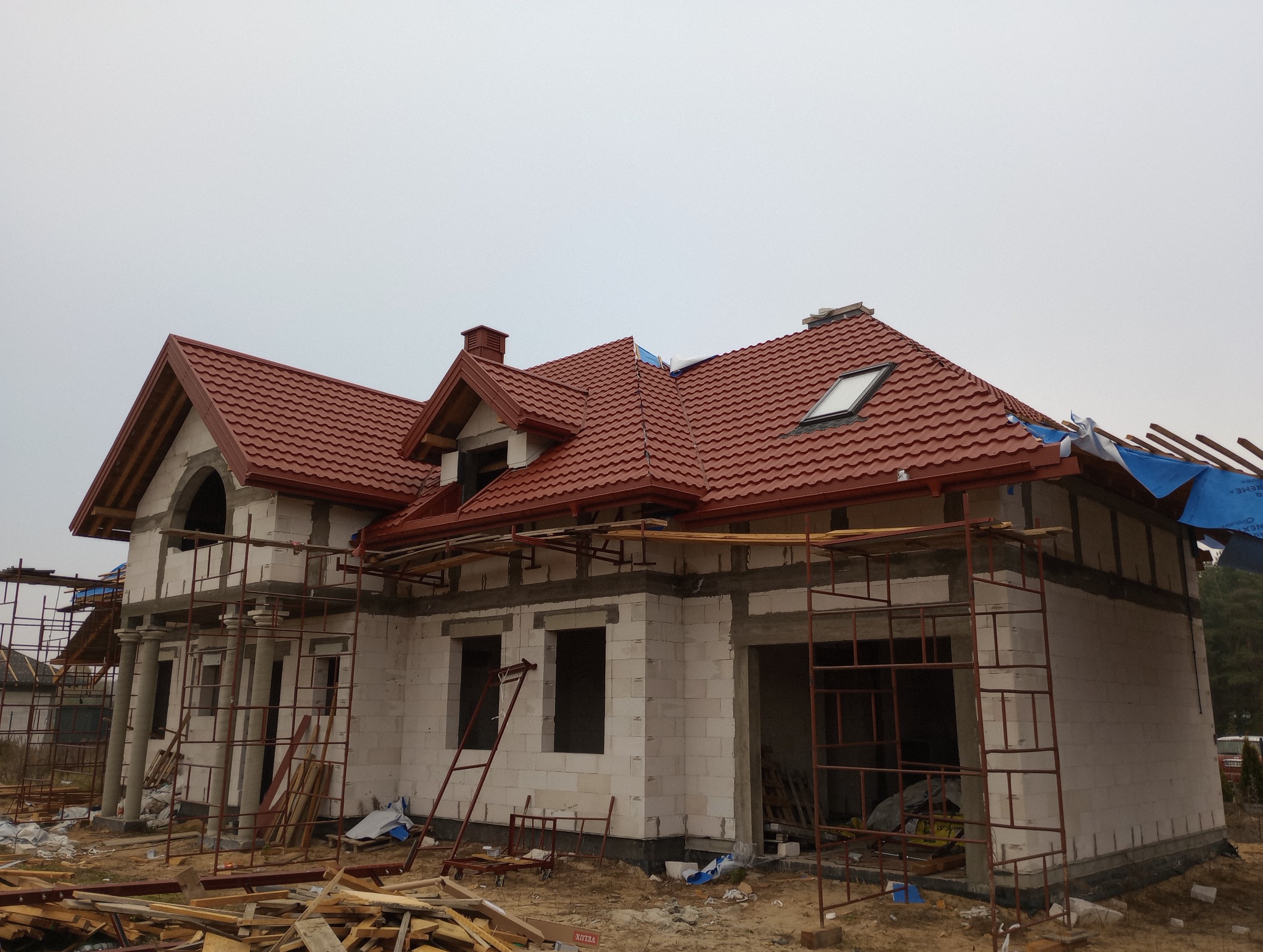 Dom w trakcie budowy z częściowo ukończonym dachem z czerwonej dachówki, widoczne rusztowania i materiały budowlane, niebo pochmurne.