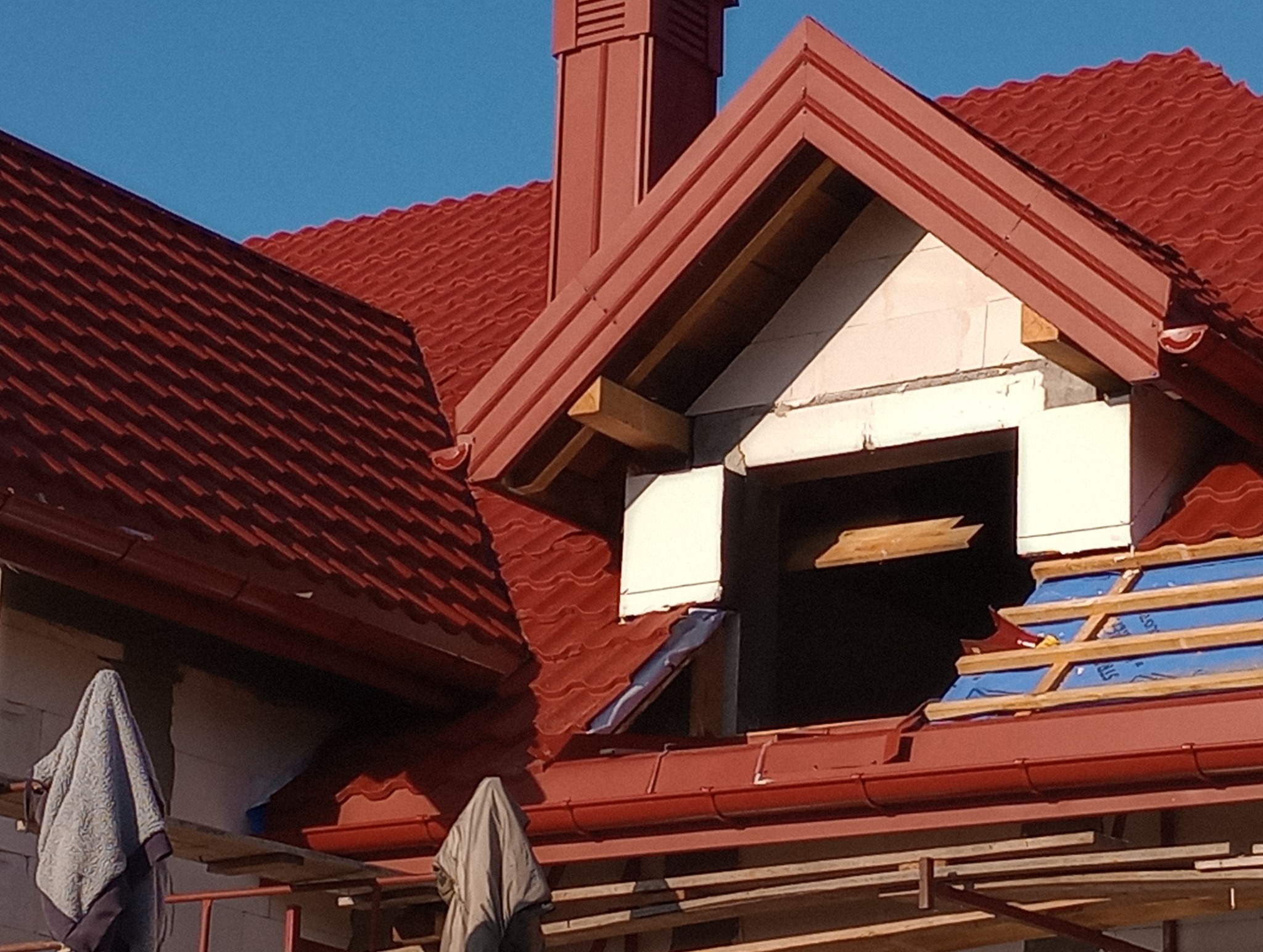 Czerwony dach z blachy trapezowej w trakcie budowy, widoczny komin, okno dachowe w stanie surowym, fragment rusztowania i płachty materiału.