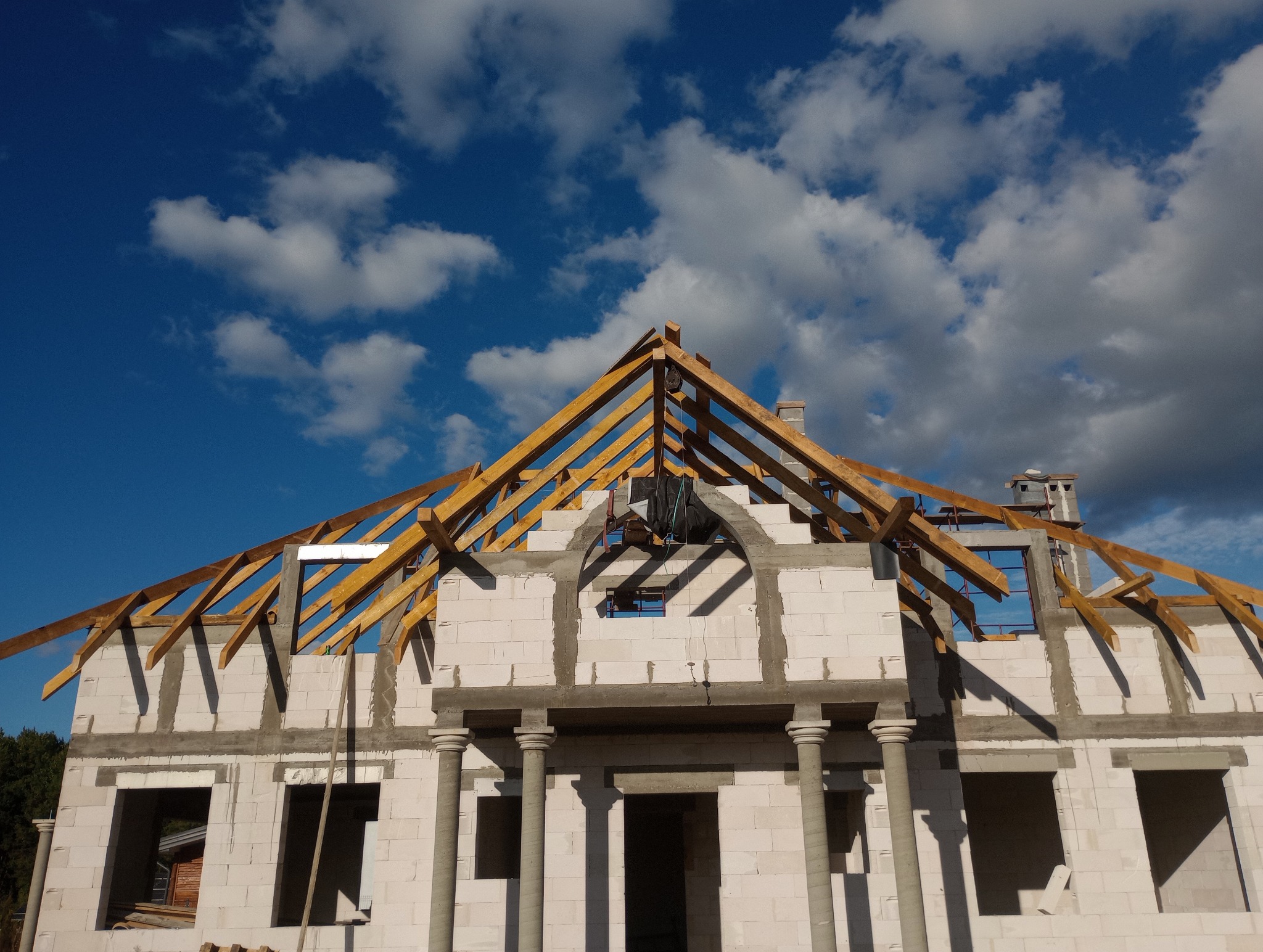 Surowy stan domu jednorodzinnego z widoczną konstrukcją drewnianą dachu na tle błękitnego nieba z chmurami.