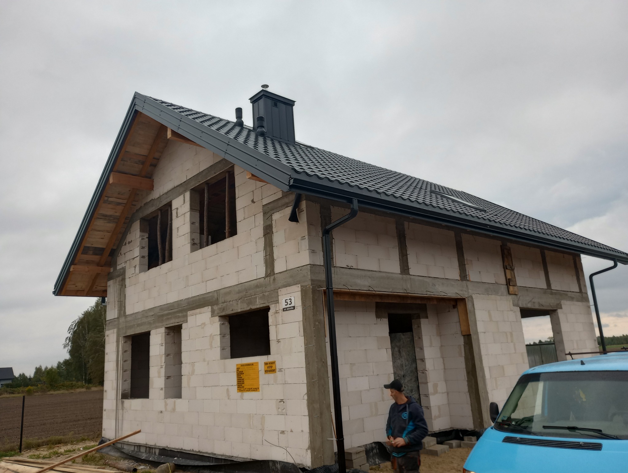 Dom w stanie surowym z zamontowanym czarnym dachem z blachodachówki i orynnowaniem; widoczny pracownik w tle i błękitny samochód dostawczy.
