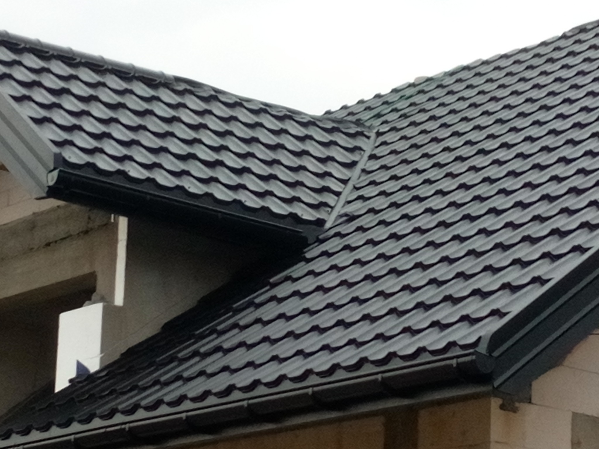 Nowoczesne, grafitowe pokrycie dachowe z blachy trapezowej, widoczne rynny, fragment ściany budynku w stanie surowym, pochmurne niebo w tle.