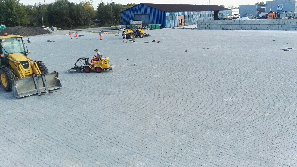 Żółta ładowarka czołowa i mała maszyna do wyrównywania kostki brukowej na rozległym placu wyłożonym szarą kostką, z pracownikami w pomarańczowych kombinezonach w tle i budynkiem przemysłowym w oddali.