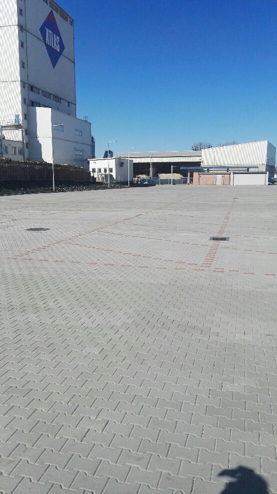 Rozległy, szary plac wyłożony kostką brukową typu 'psi kość', z widocznymi liniami z czerwonej kostki, w tle budynki przemysłowe i błękitne niebo.