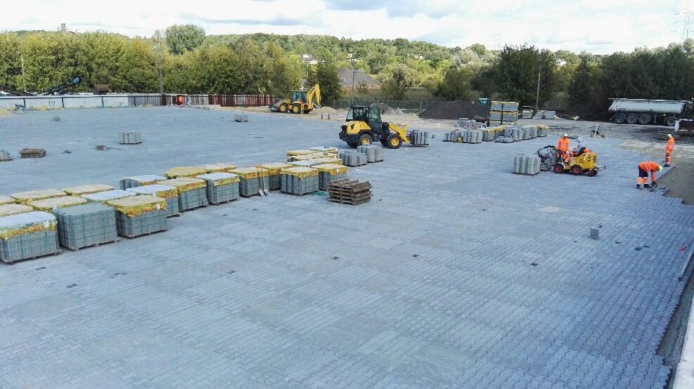 Rozległy plac z częściowo ułożoną szarą kostką brukową, palety z kostką w żółtych osłonach, koparka i ładowarka w tle, pracownicy w pomarańczowych kombinezonach przy układaniu kostki.