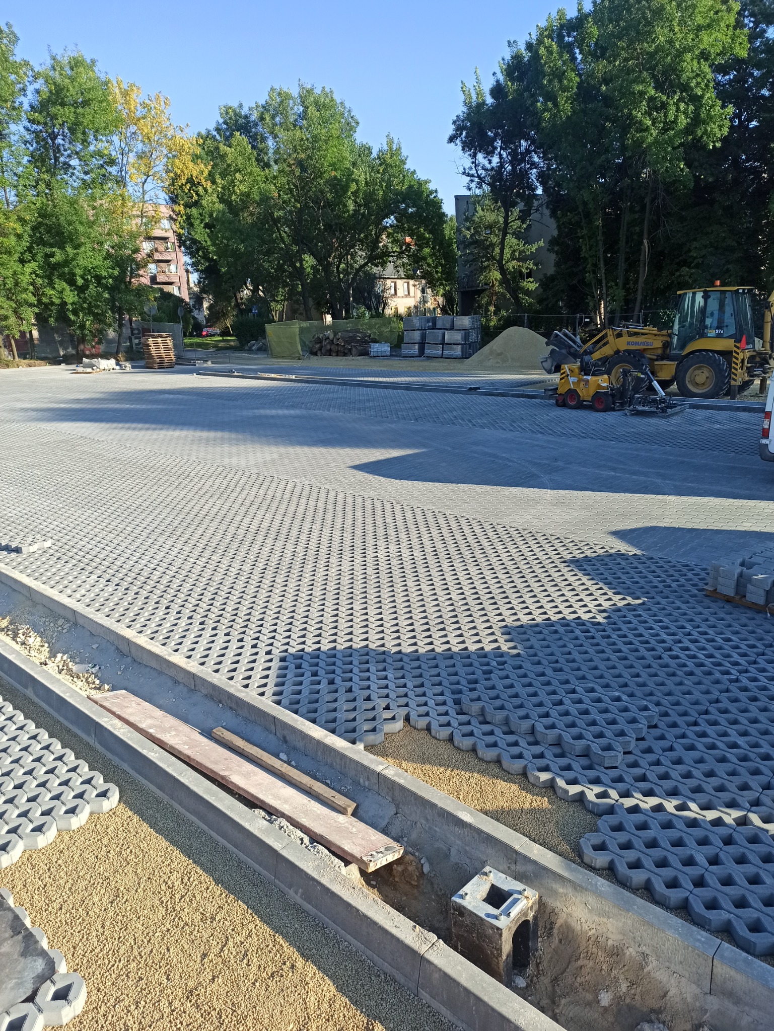 Układanie ażurowej kostki betonowej na podsypce piaskowej, widoczny fragment korytka odwadniającego z metalową obudową, w tle maszyny budowlane i drzewa.