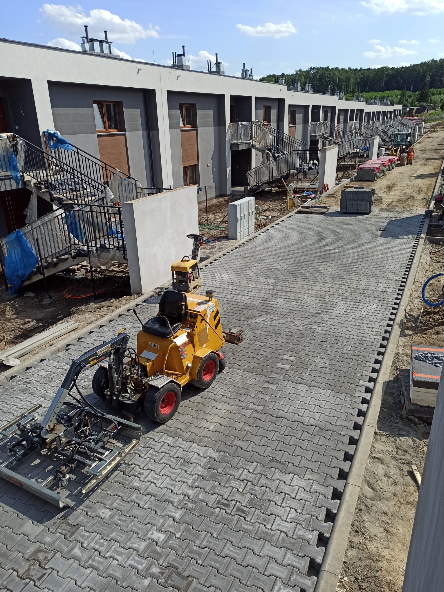 Żółta maszyna do układania kostki brukowej na szarym brukowanym chodniku przed szeregiem nowych domów w trakcie budowy, z paletami kostki w tle.