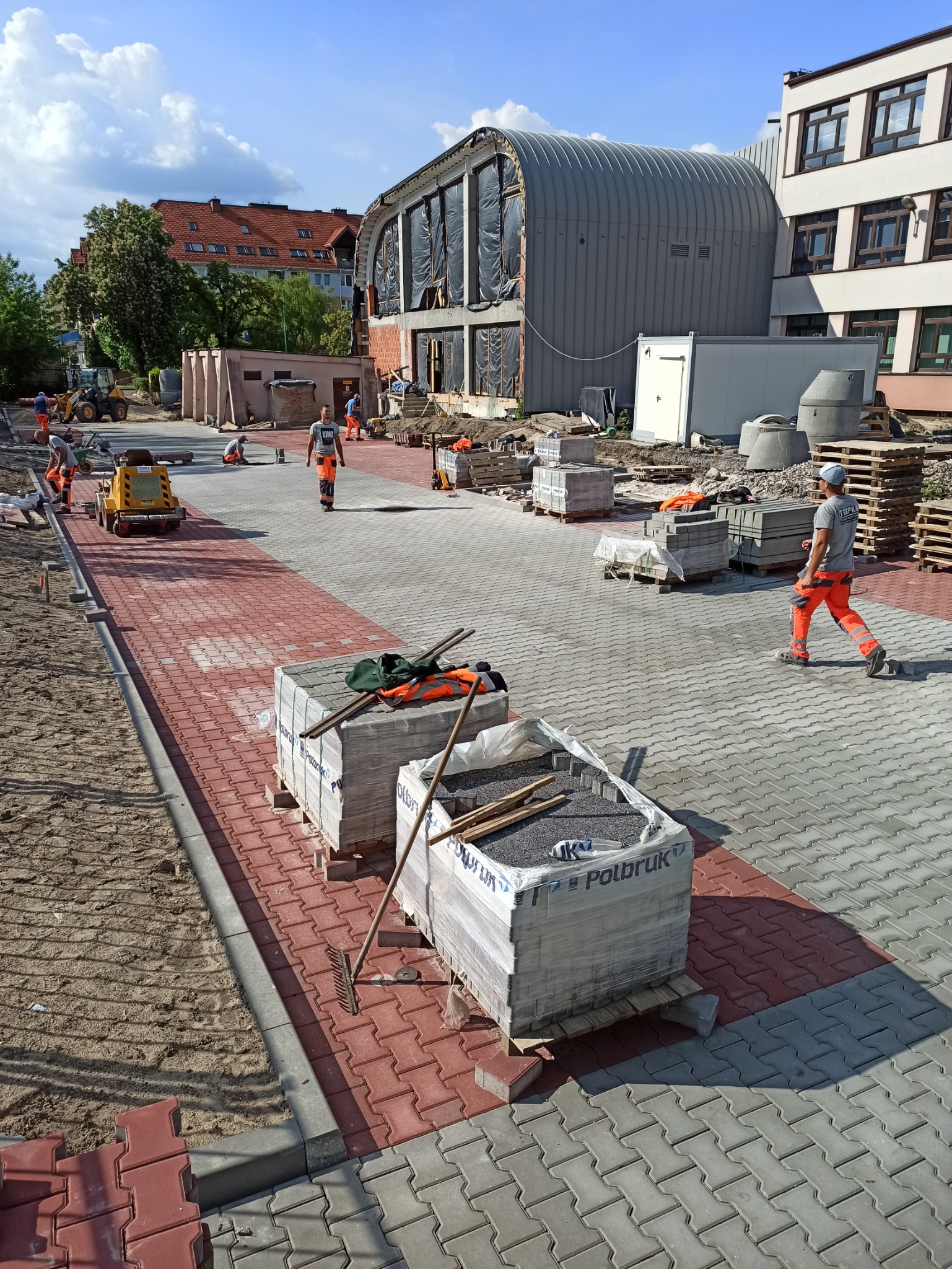 Trwa układanie dwukolorowej kostki brukowej (czerwono-szarej) na placu budowy. Widoczne palety z kostką Polbruk, narzędzia, pracownicy w pomarańczowych kombinezonach oraz maszyna do ubijania. W tle...