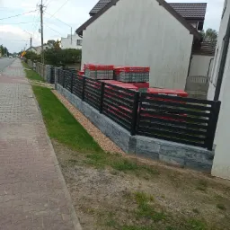 Nowoczesne, czarne ogrodzenie panelowe na podmurówce z szarej kostki, obok chodnika z czerwonej kostki brukowej, z paletami czerwonej kostki w tle.