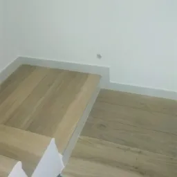 Fragment podłogi wykończonej panelami imitującymi drewno, z widoczną białą listwą przypodłogową i fragmentem szablonu malarskiego.