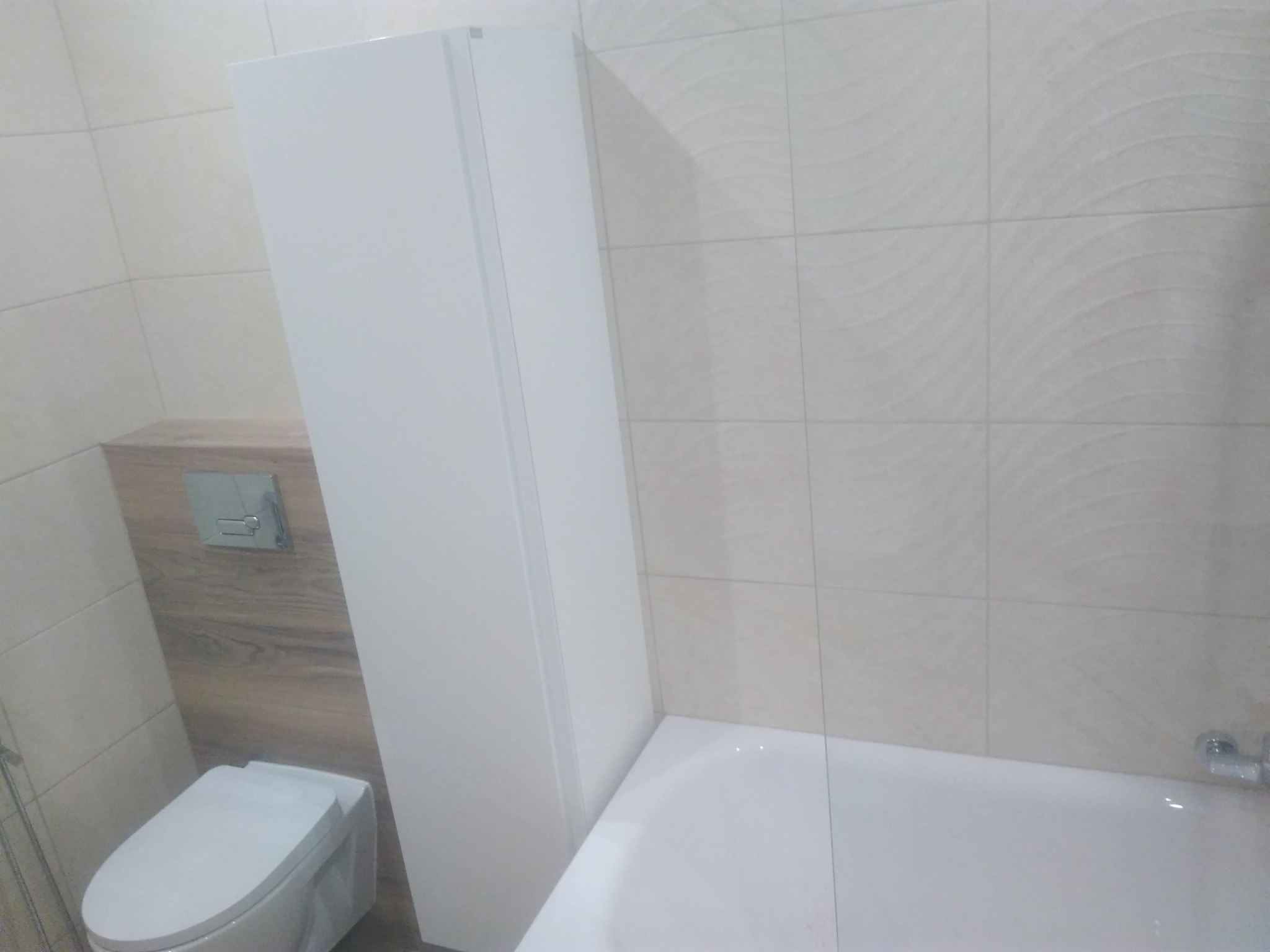 Nowoczesna łazienka z beżowymi płytkami, zabudowaną toaletą z drewnianym panelem i białą szafką wiszącą obok wanny.